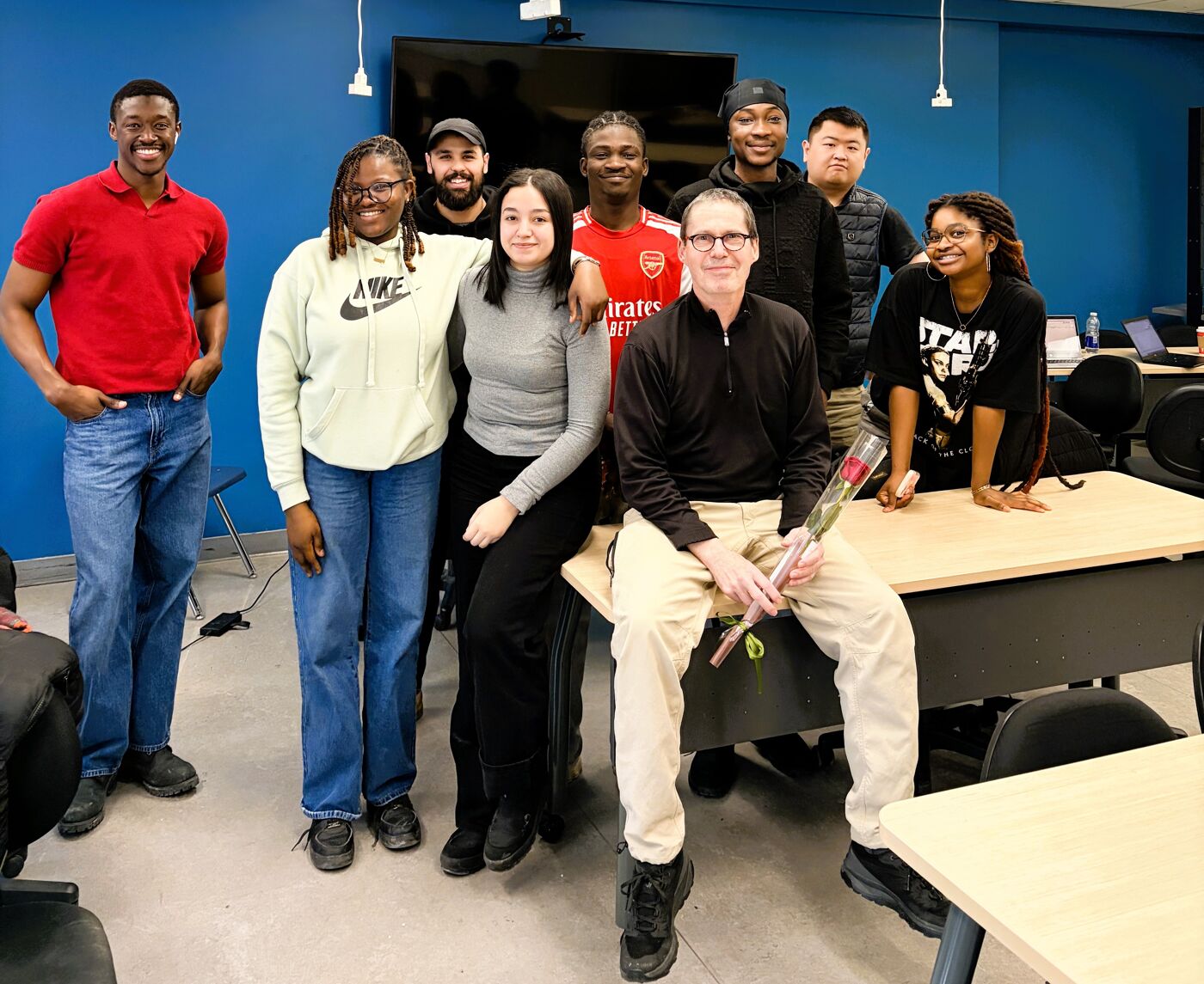 A group of diverse people are posing for a photo in a classroom setting. Some are smiling, and one person is holding a rose.