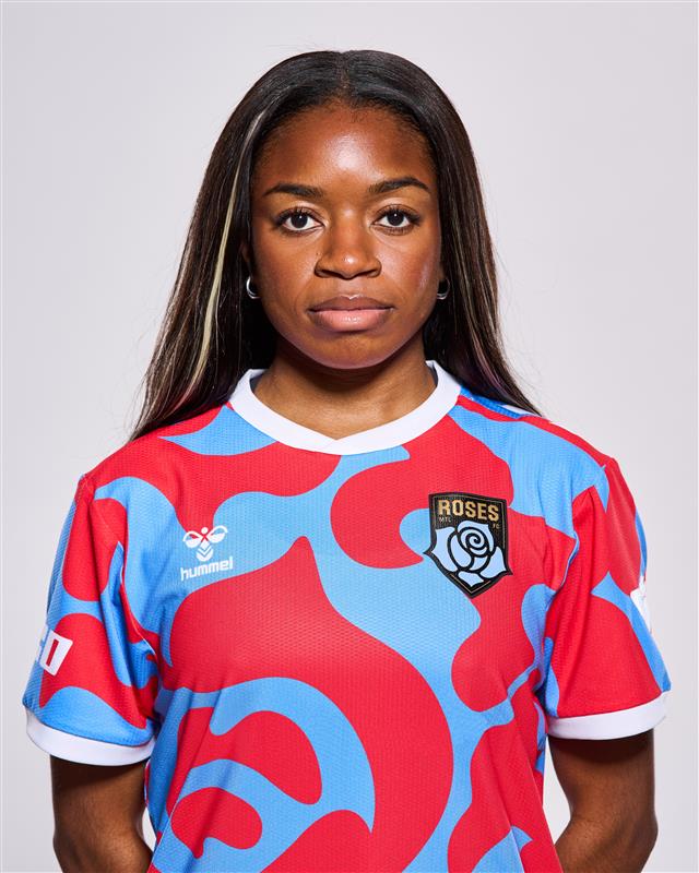 A young woman with dark skin is wearing a red and blue soccer jersey with a Roses logo on the left sleeve.