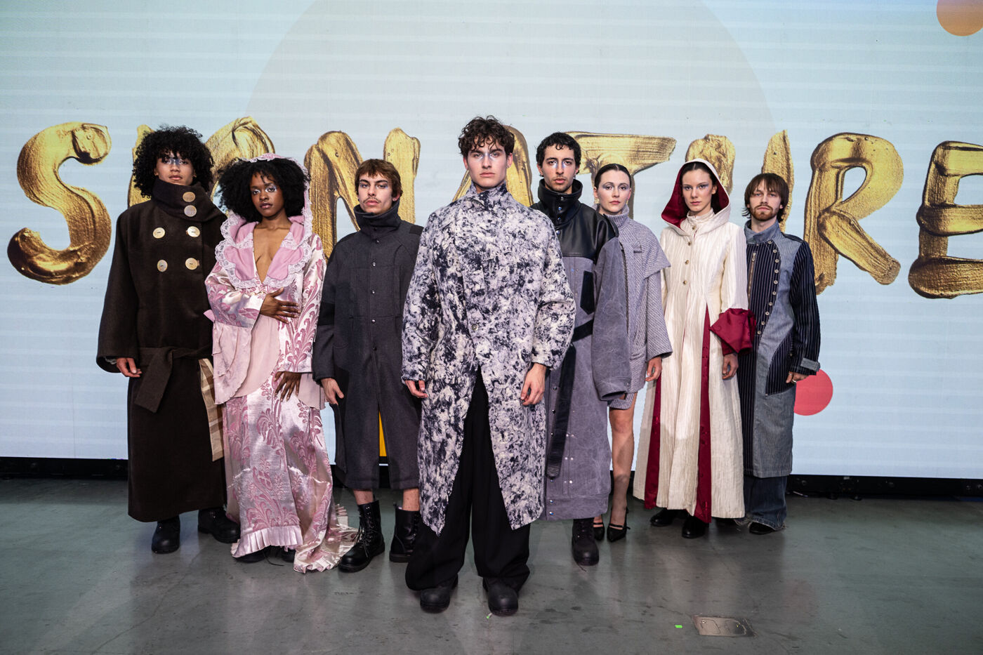 A group of nine individuals, both men and women, stand before a backdrop featuring the text 'SAINT LAURENT' in golden 3D lettering. They are all dressed in unique and stylish garments, showcasing diverse textures and patterns, suggesting a fashion show.