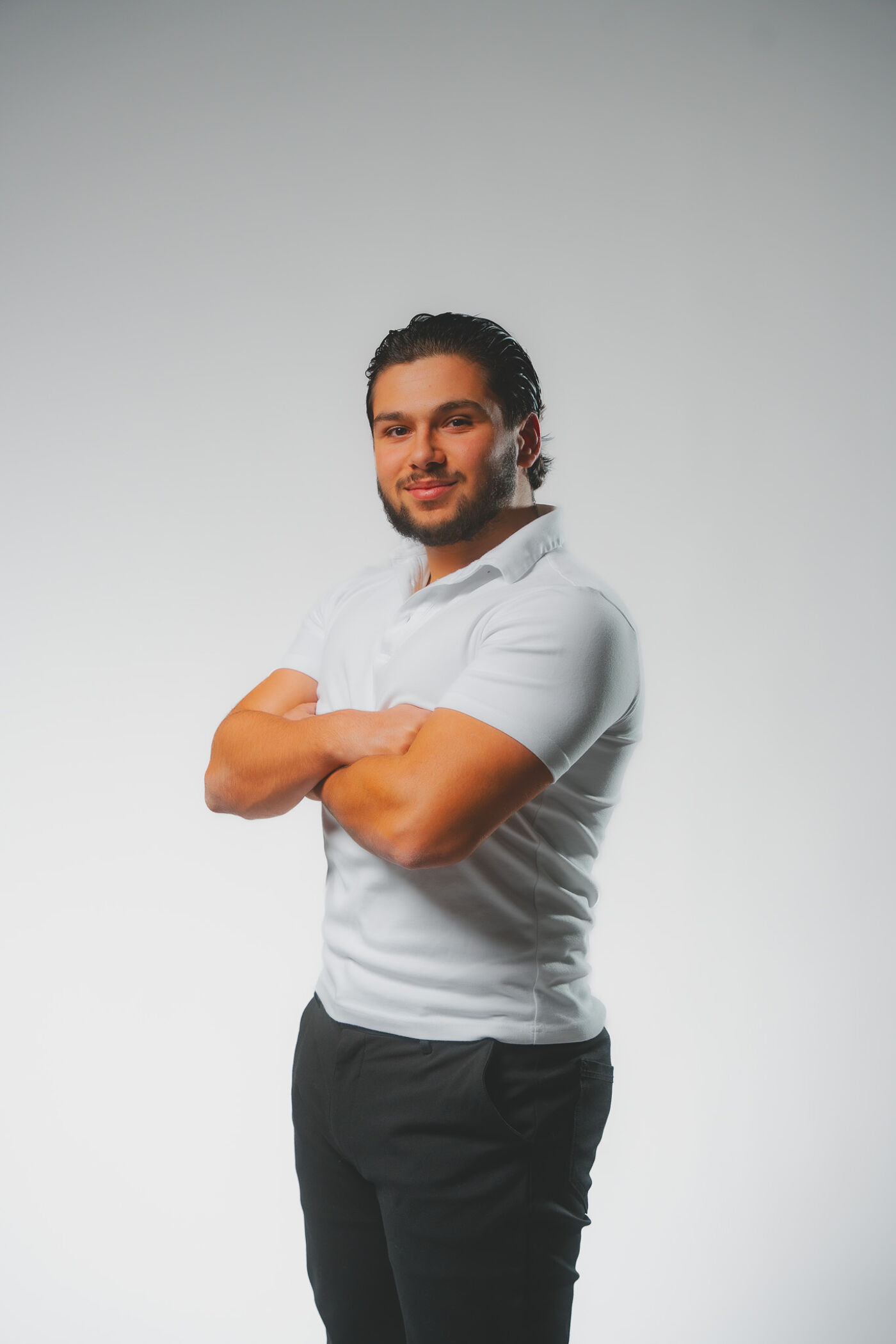 A young man with crossed arms, wearing a white polo shirt and black pants, looks towards the camera against a gray background.