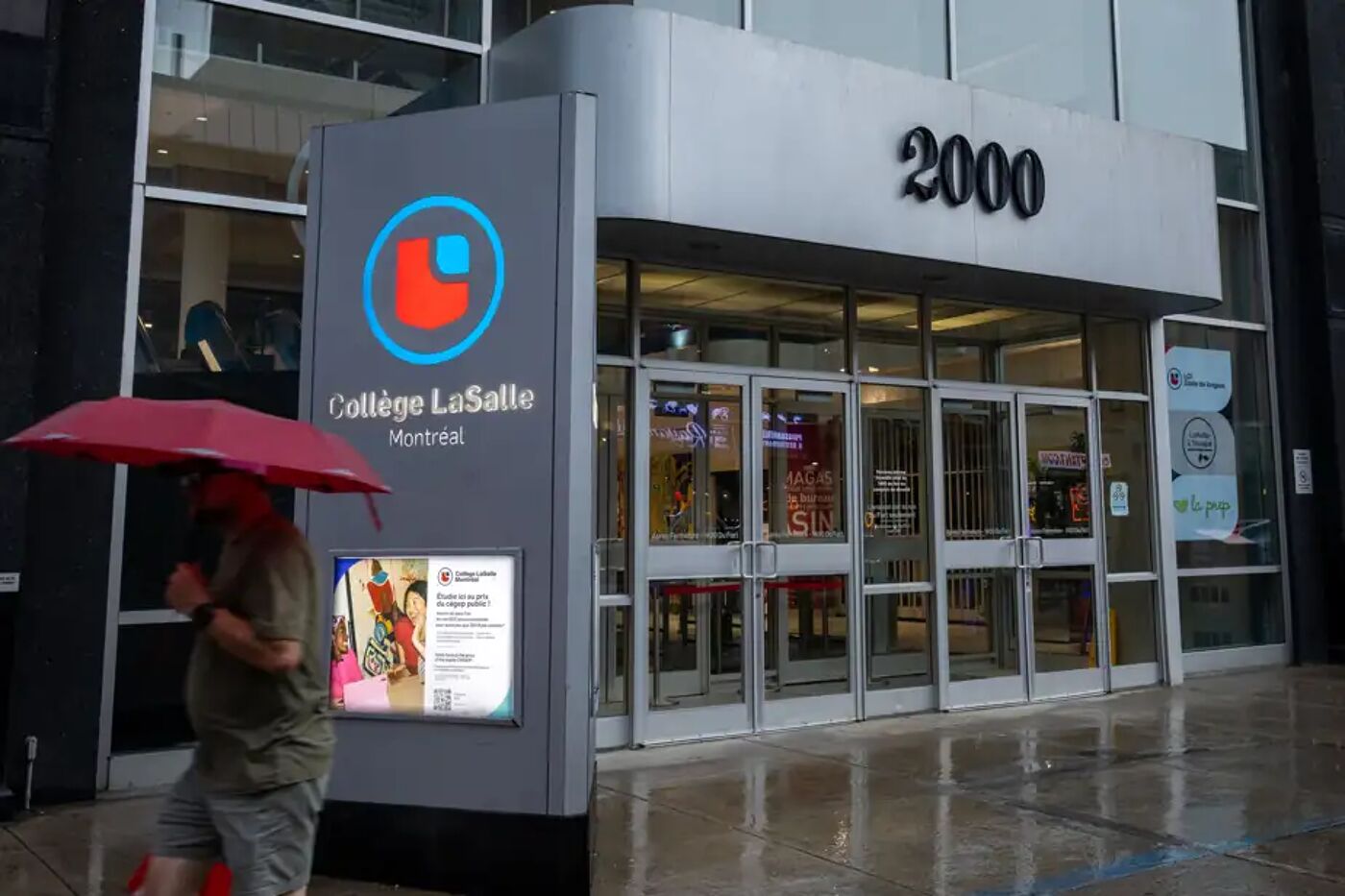 A person holding a red umbrella walks past the entrance of Collège LaSalle in Montreal, with the number 2000 above the doors.