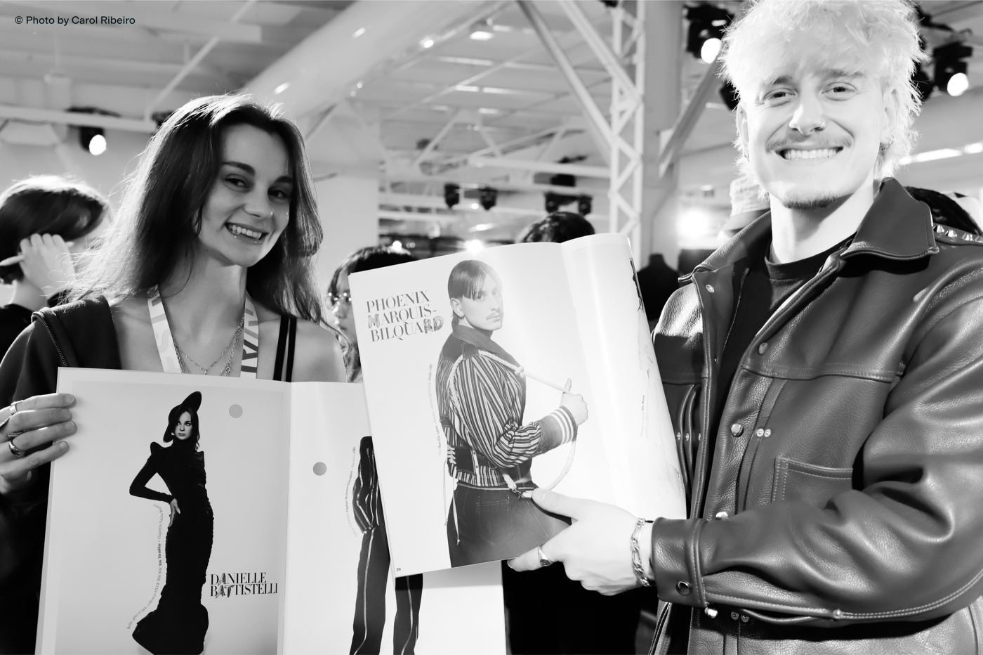 Two people smile at the camera while holding an open book featuring fashion images, including a woman in an elegant dress and a man in a striped sweater.