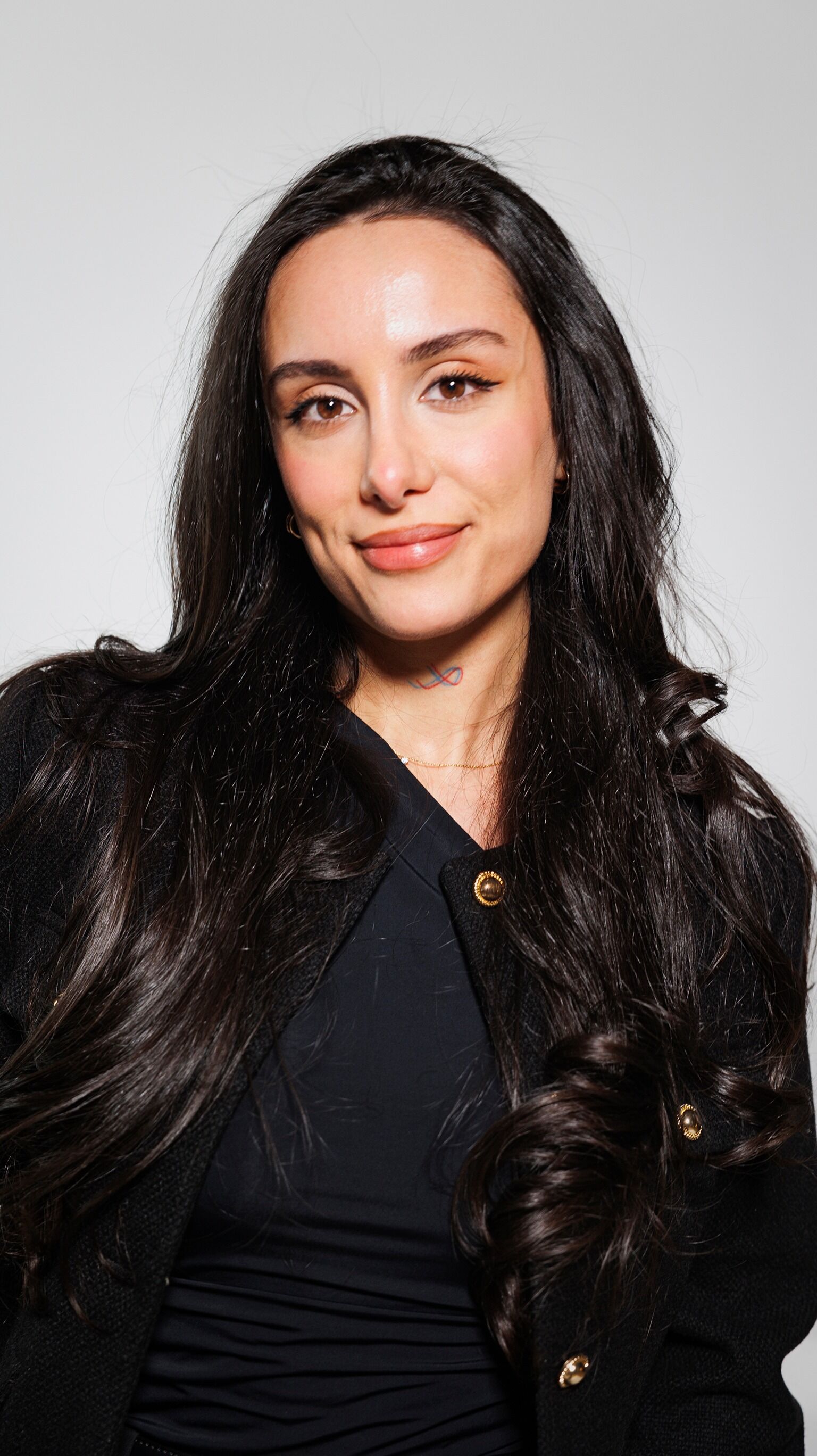 The image shows a woman with long, dark hair and a black outfit. She is smiling at the camera, and the background is plain.