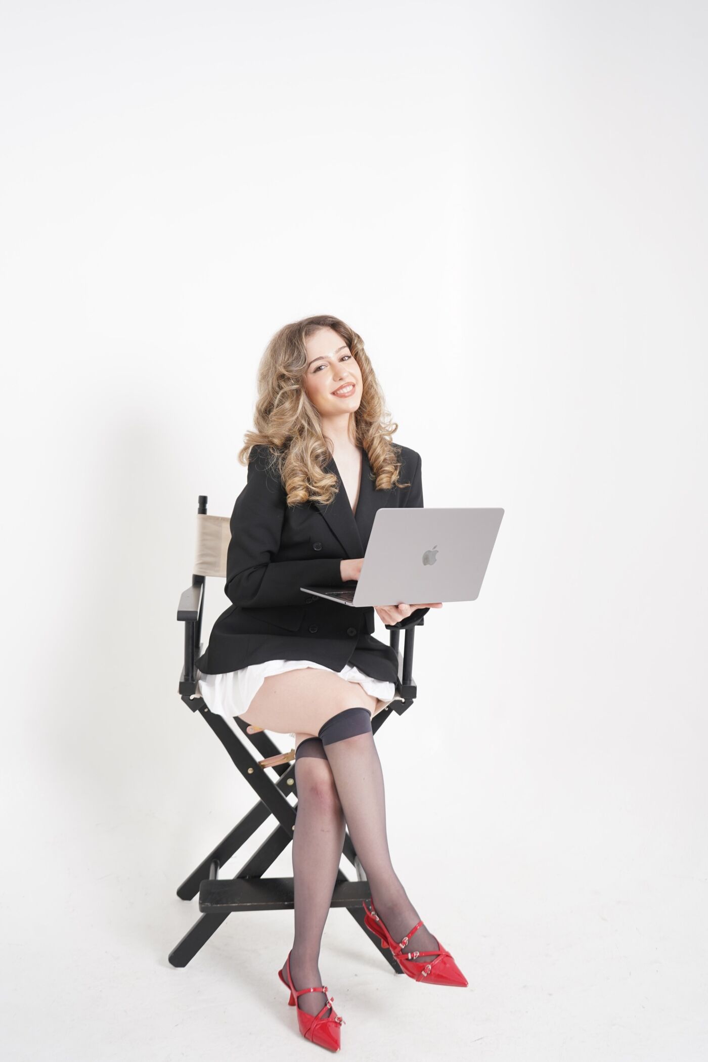 A woman with curly hair is sitting on a director's chair, holding a laptop. She is wearing a black blazer, a white skirt, and red shoes.