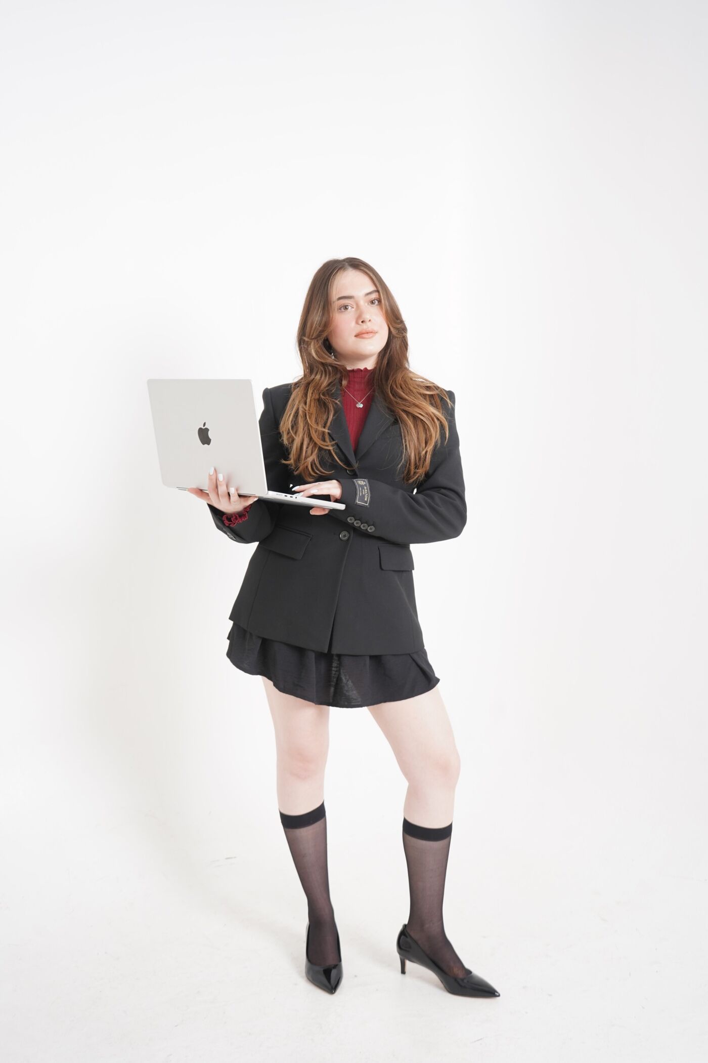 The image shows a woman in a black suit holding a laptop. She is wearing a red shirt, a black skirt, and black stockings. She is standing in front of a white background.