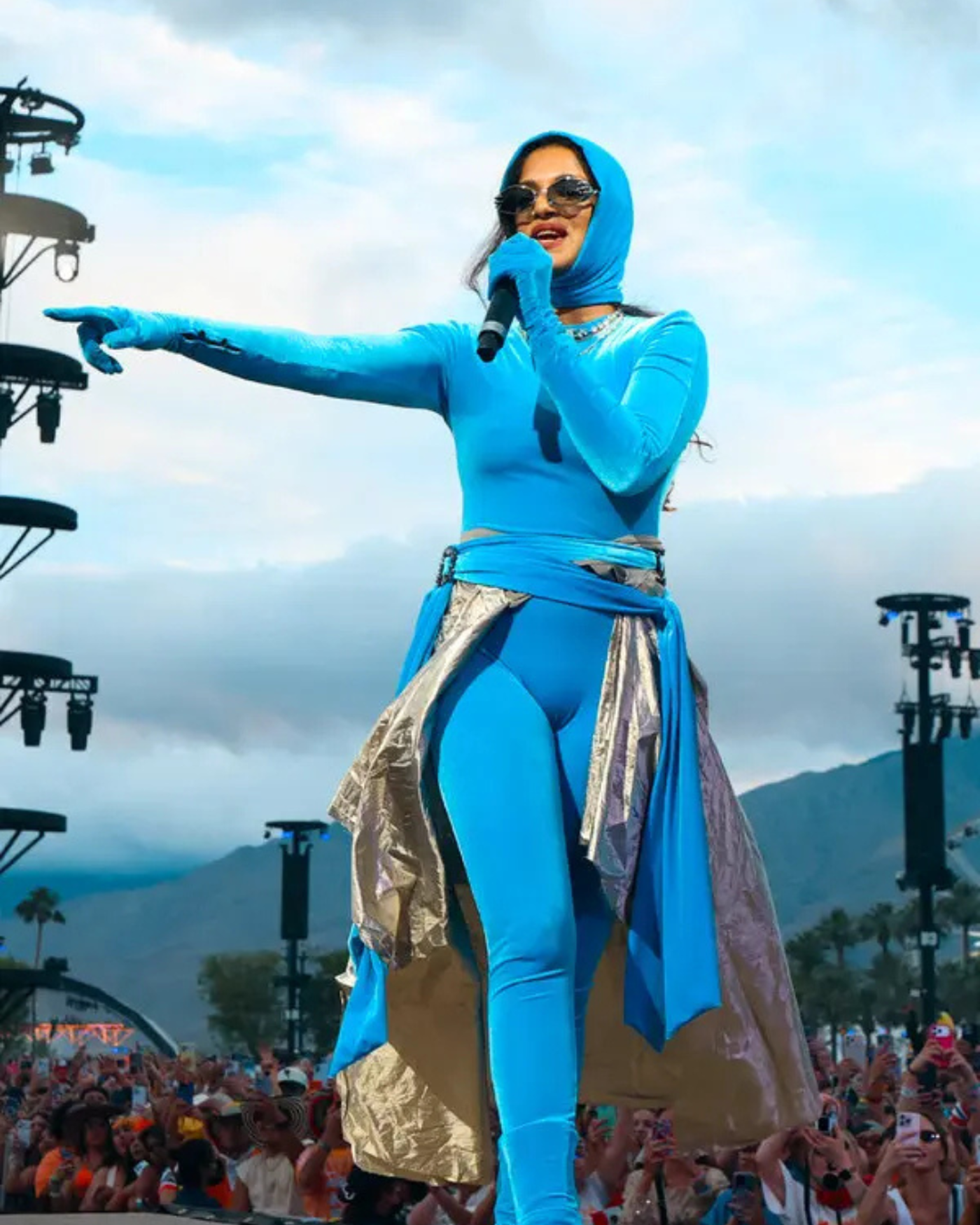 A singer in a vibrant blue outfit, complete with a hood, performs on stage before a crowd. She wears sunglasses and a necklace, with a silver and gold skirt over her fitted pants.