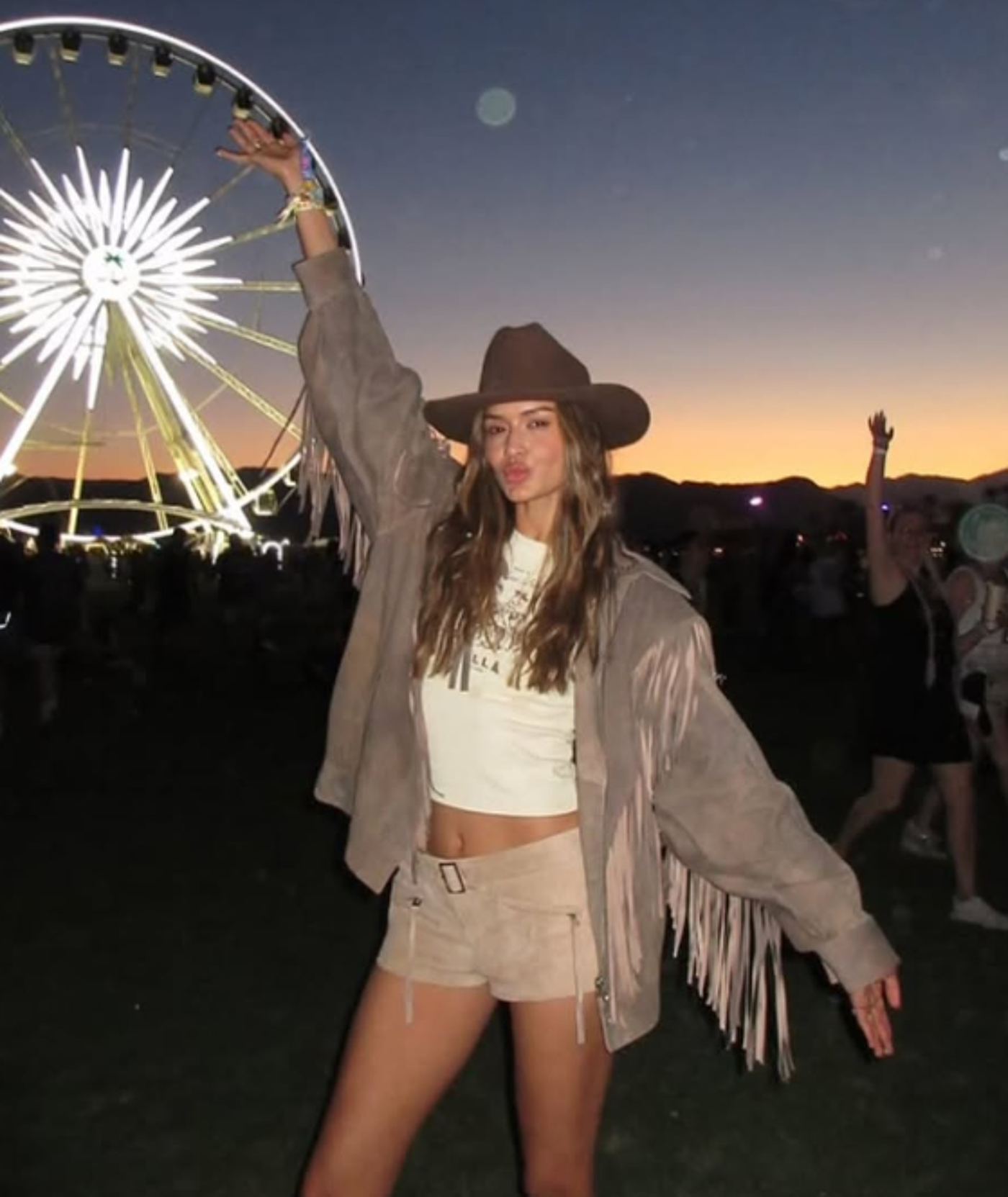 A young woman with long brown hair, wearing a brown fringed jacket, beige shorts, and a brown cowboy hat, poses with one arm raised in front of a Ferris wheel at sunset.