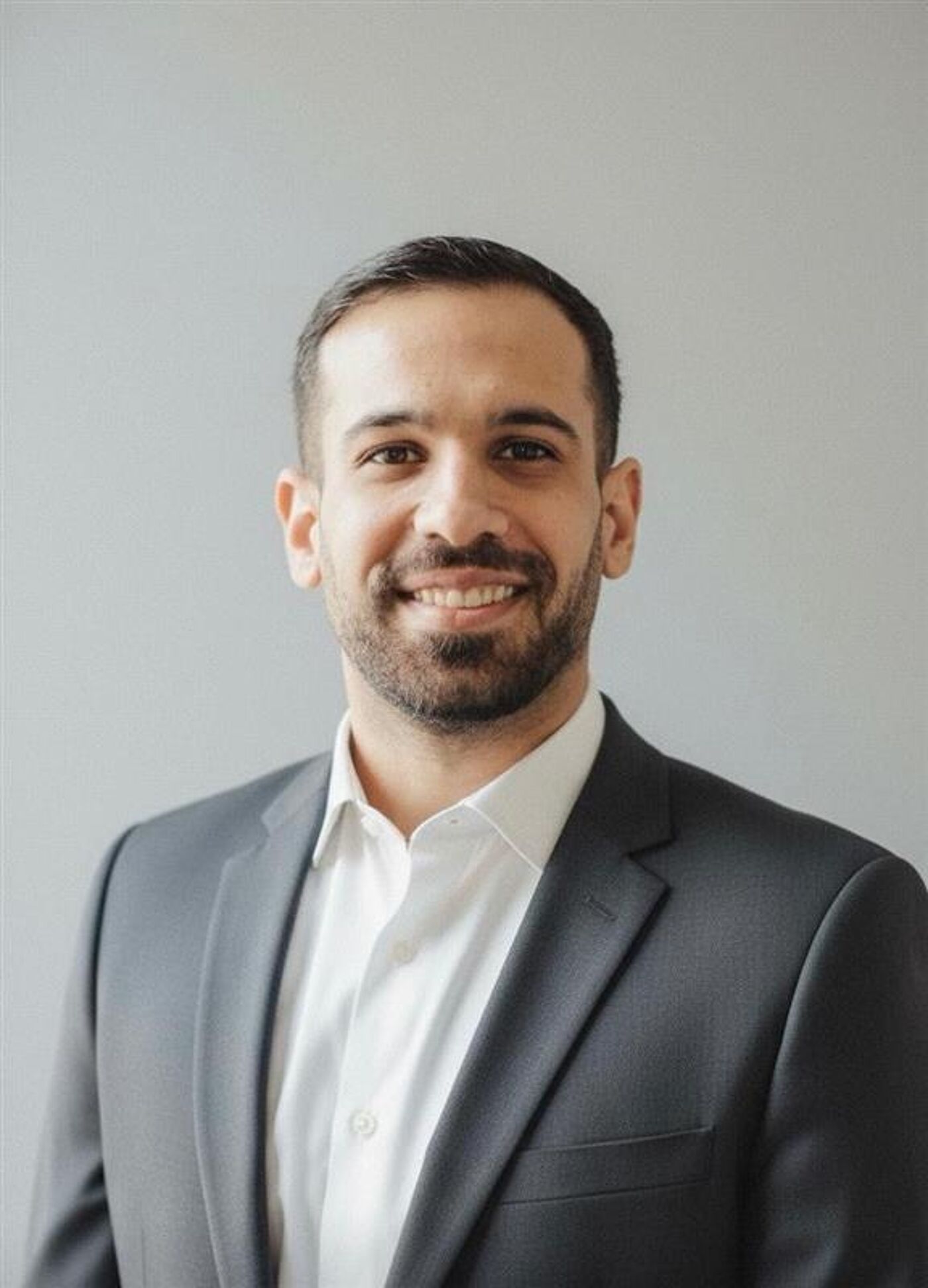 This is a close-up photograph of a smiling man wearing a grey suit and a white shirt. He has short, dark hair and a short beard.