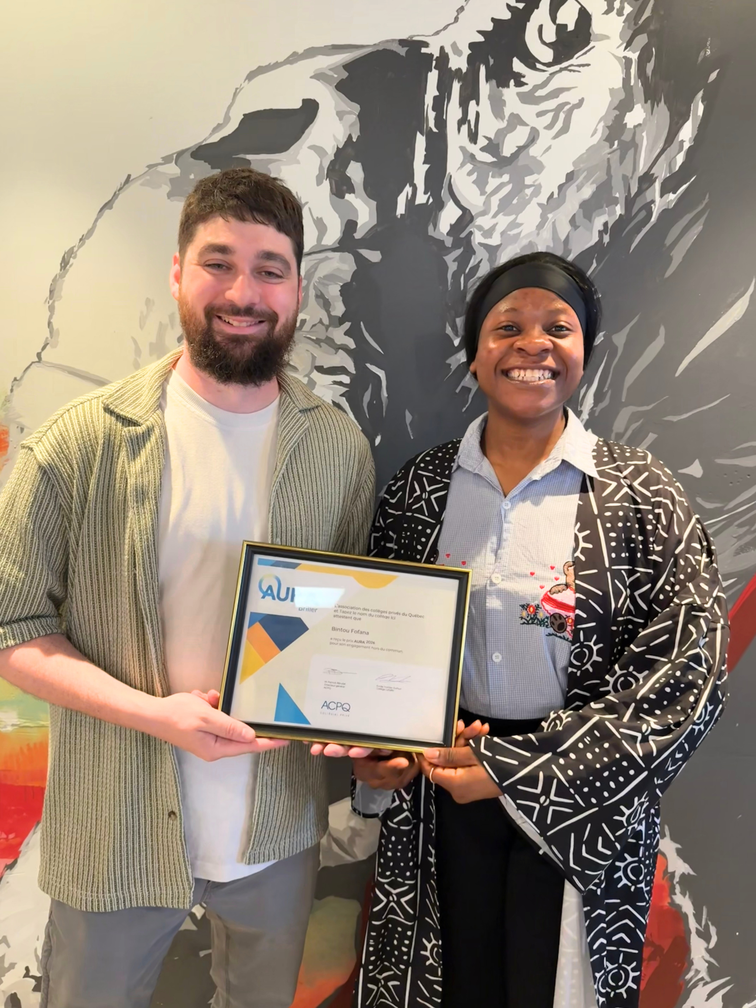 Two people are holding a certificate with the WUI logo. It appears to be an award or recognition, presented in front of an artistic mural.