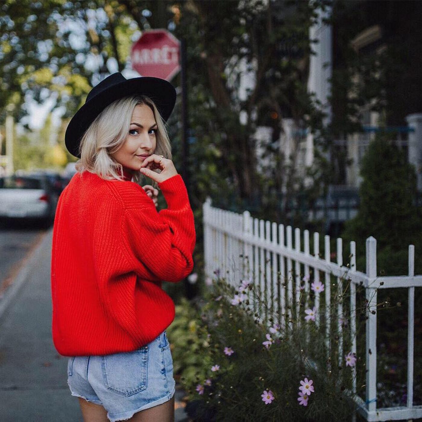 A blonde woman in a red sweater and denim shorts, wearing a black hat, stands sideways, looking over her shoulder, in front of a white picket fence with flowers.