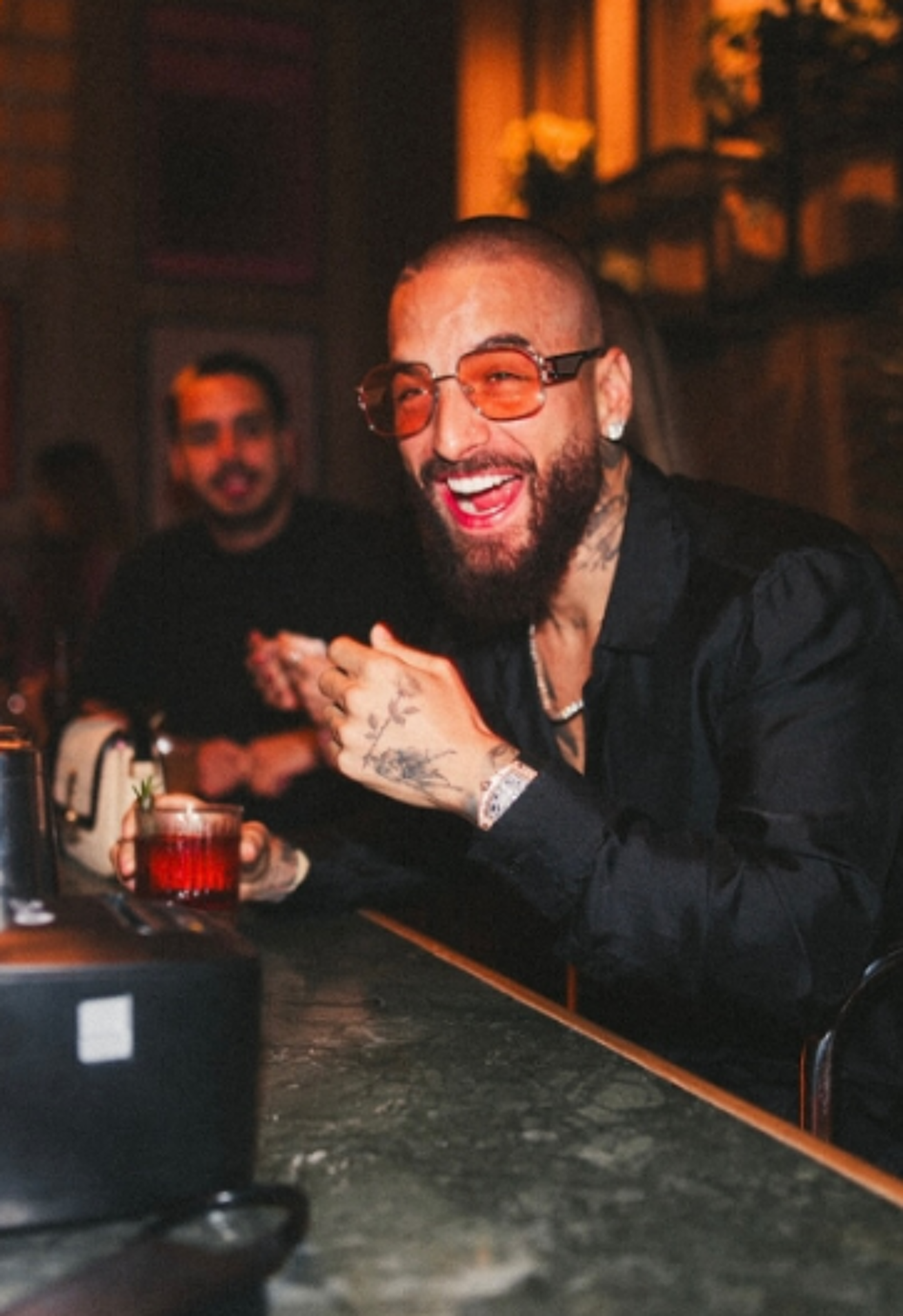 A man with orange-tinted glasses and a beard laughs while holding a cocktail at a bar, with another man blurred in the background.