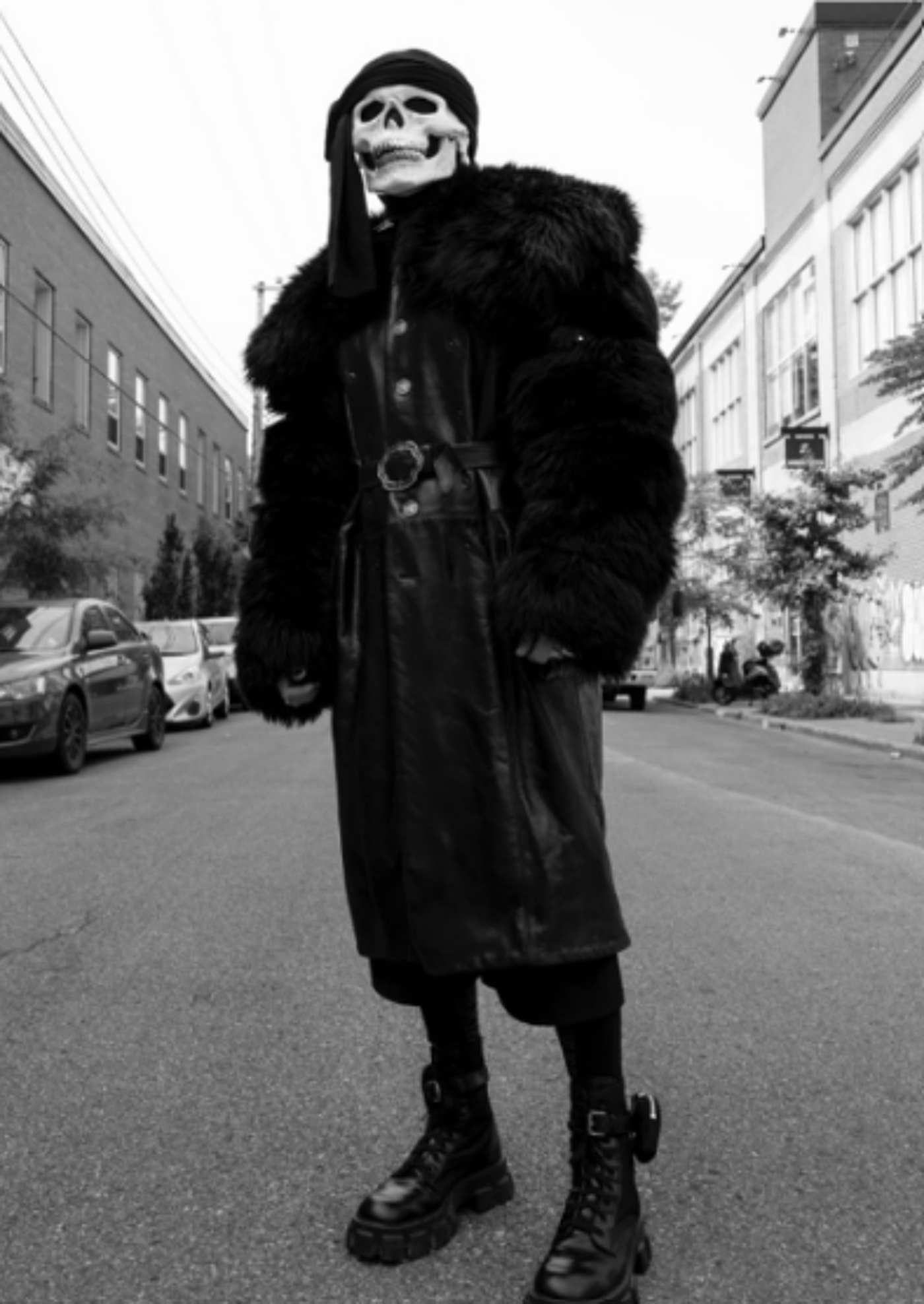 A person wearing a skull mask and long black coat stands on a city street.