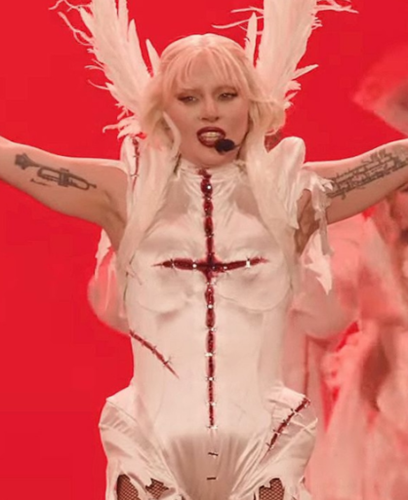 Lady Gaga, wearing a white, ripped bodysuit adorned with a red cross and large white feathered wings, sings into a microphone against a red background.