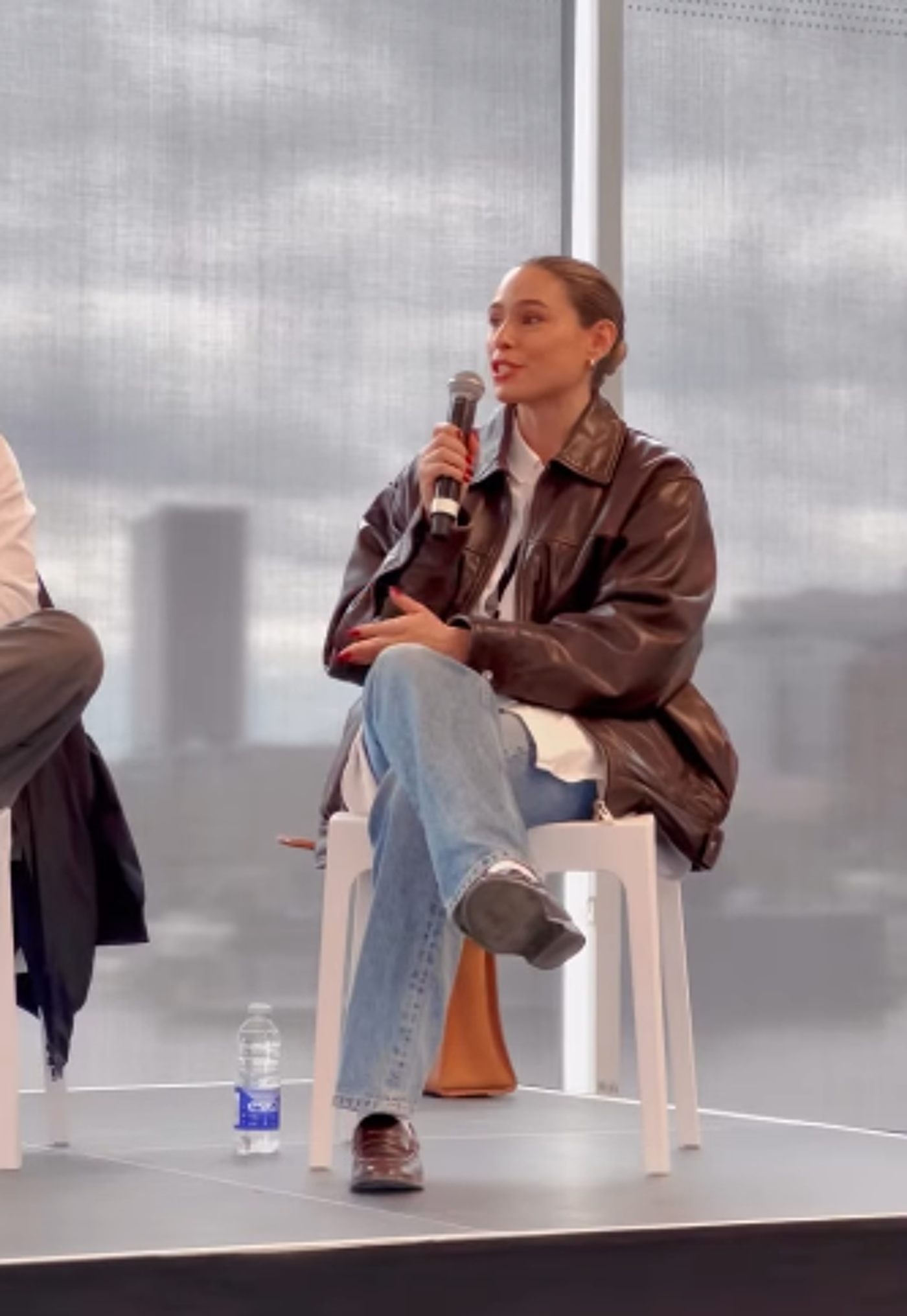 A woman in a brown leather jacket and blue jeans sits on a white chair, holding a microphone and speaking.