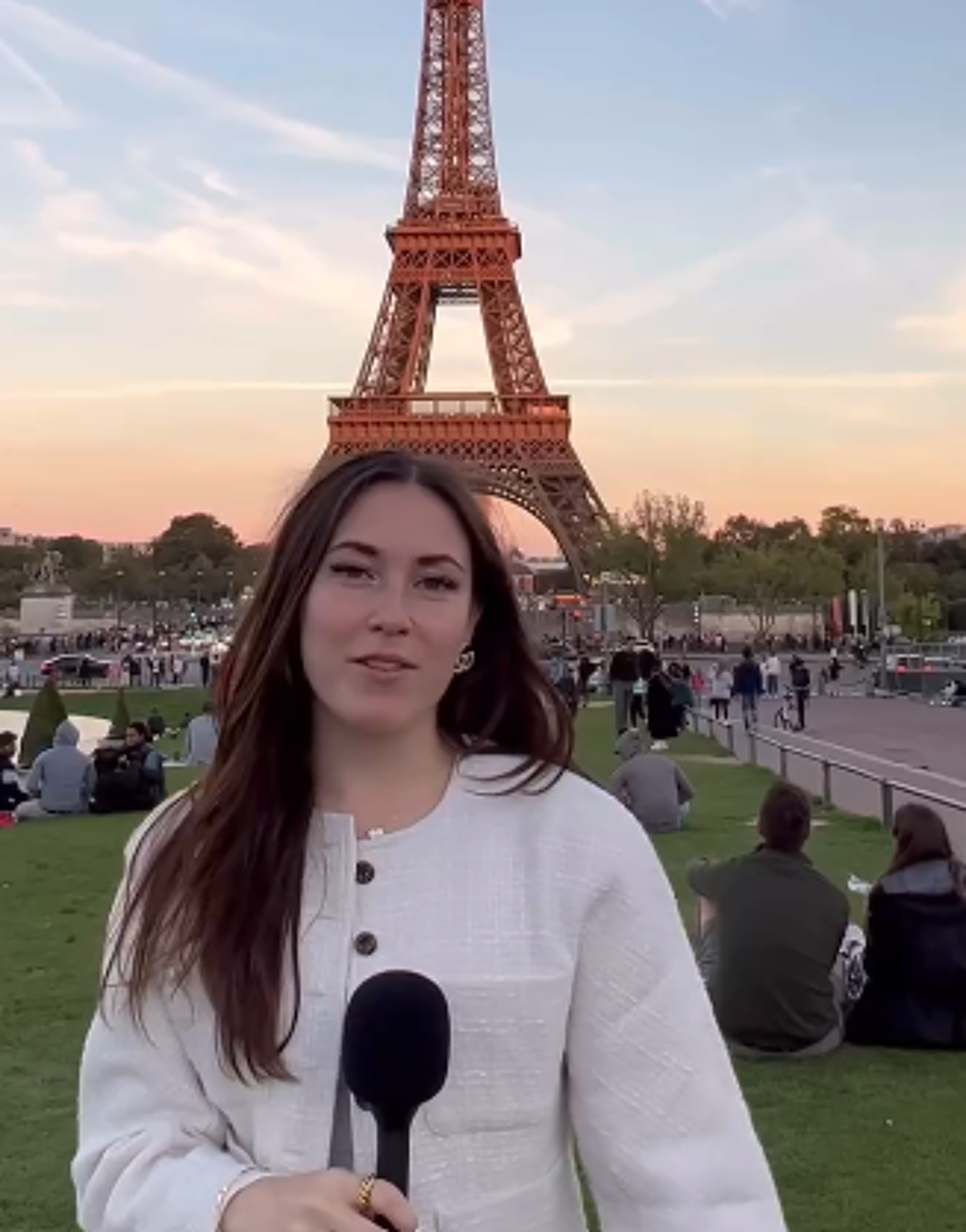 A young woman with long brown hair holds a microphone in front of the Eiffel Tower at sunset, with people relaxing on the grass in the foreground.