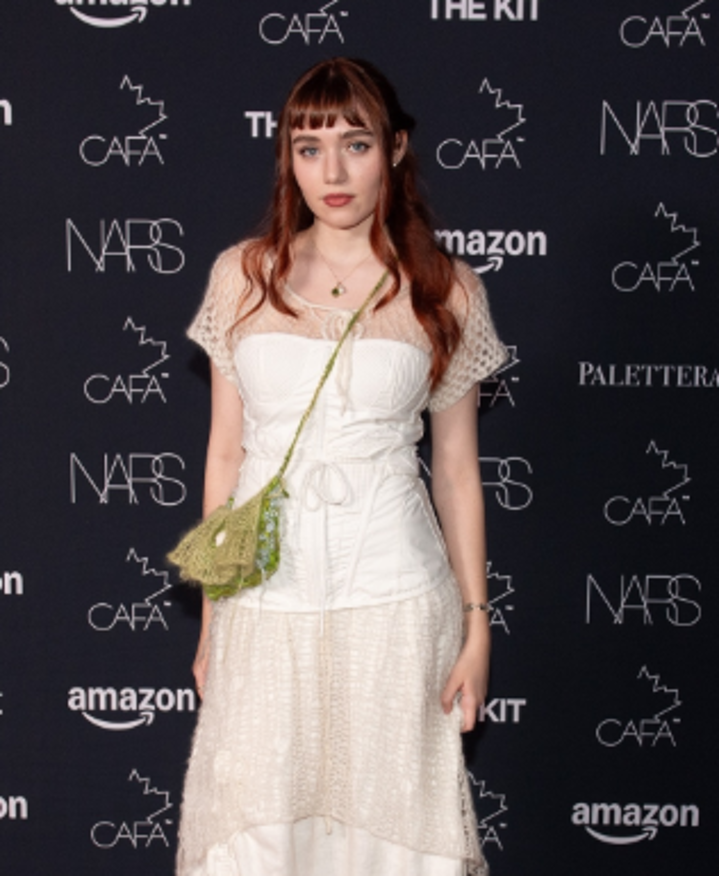 A young woman with auburn hair and bangs stands before a black backdrop adorned with various logos, wearing a white crocheted dress and a small green purse.