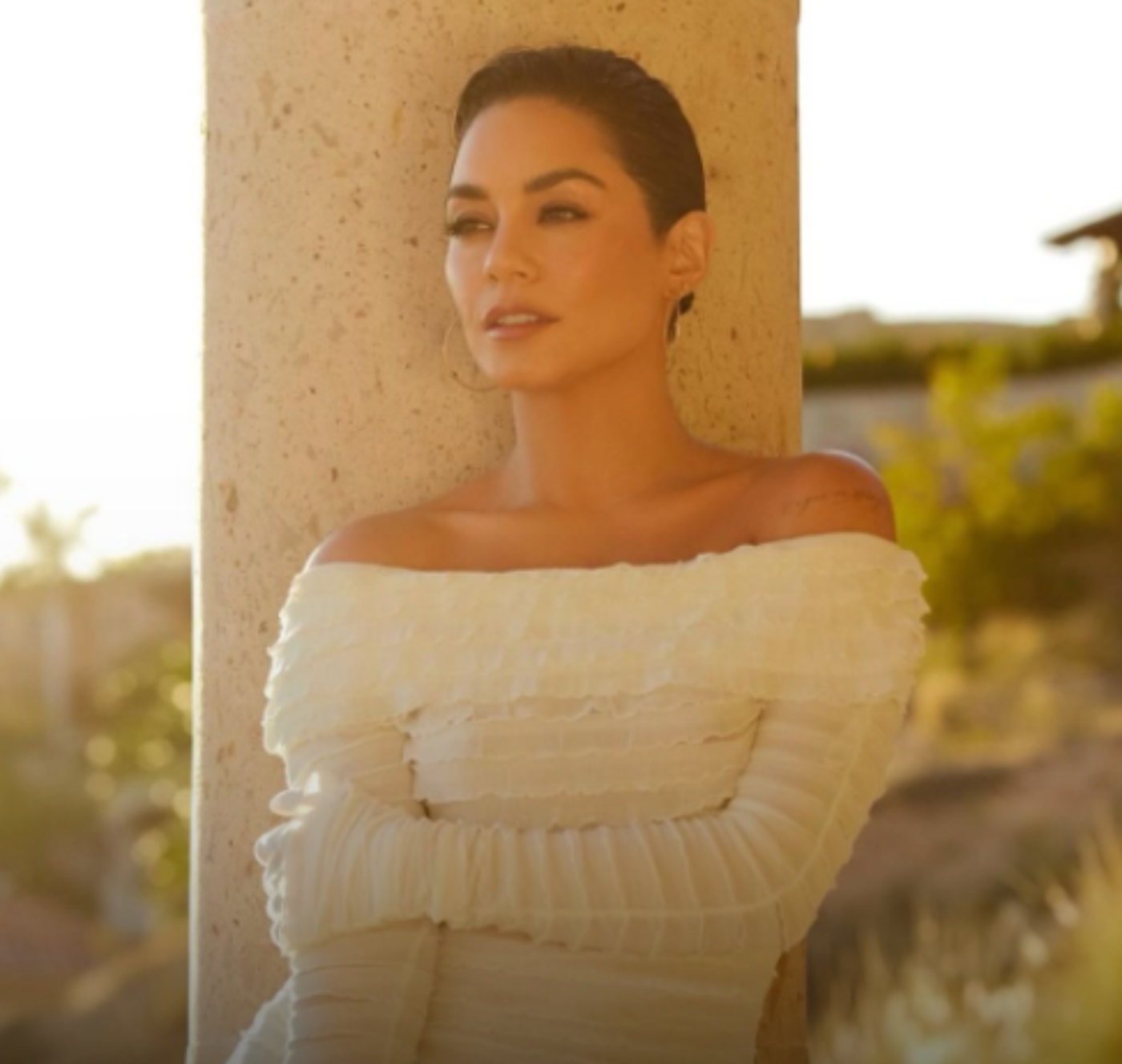 Vanessa Hudgens, with short dark hair and wearing an off-the-shoulder white ruffled top, leans against a light beige column, gazing off to her right.