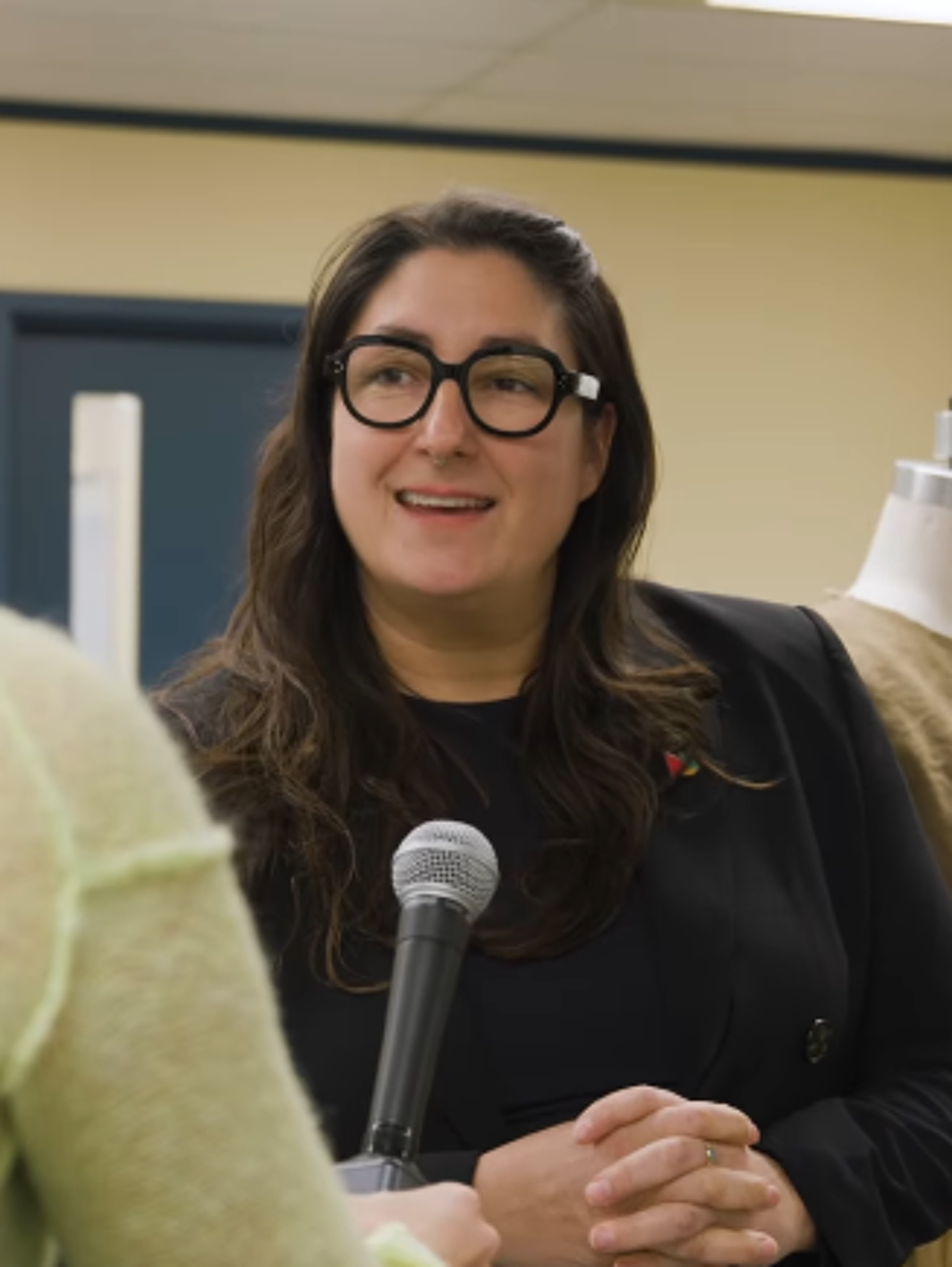 A woman with long brown hair and glasses sits with her hands clasped, looking slightly to her right, while holding a microphone.