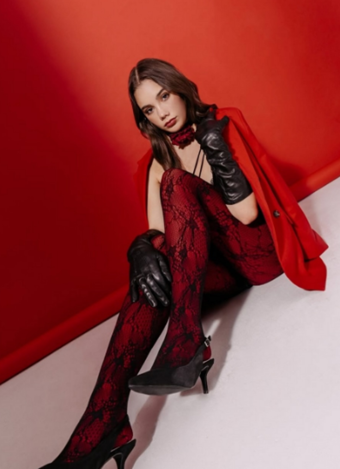 A woman in red lace tights and a red blazer sits on a red background, wearing black leather gloves and heels.