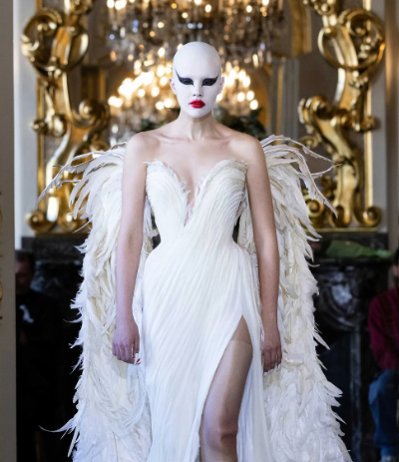A bald model with dramatic makeup and large feathered wings wears a white, figure-hugging gown with a thigh-high slit at a fashion show.