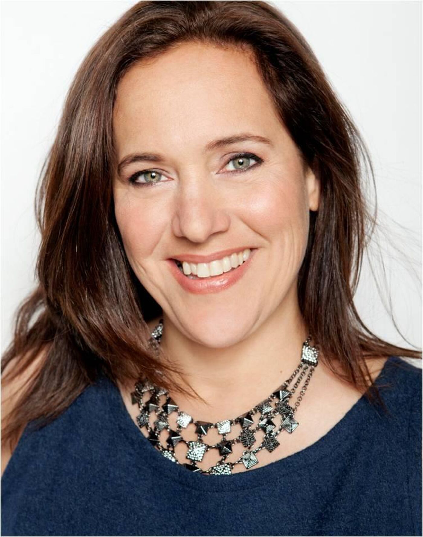 Close-up shot of a woman with shoulder-length brown hair, wearing a dark blue top and a layered necklace with square and pyramid-shaped accents.