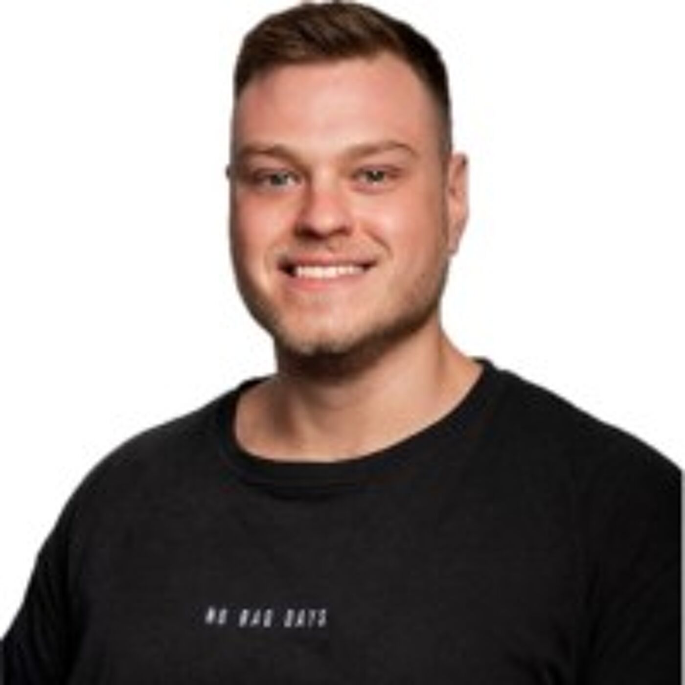 Close-up shot of a smiling, light-skinned man with short brown hair, wearing a black crew-neck t-shirt with subtle text.