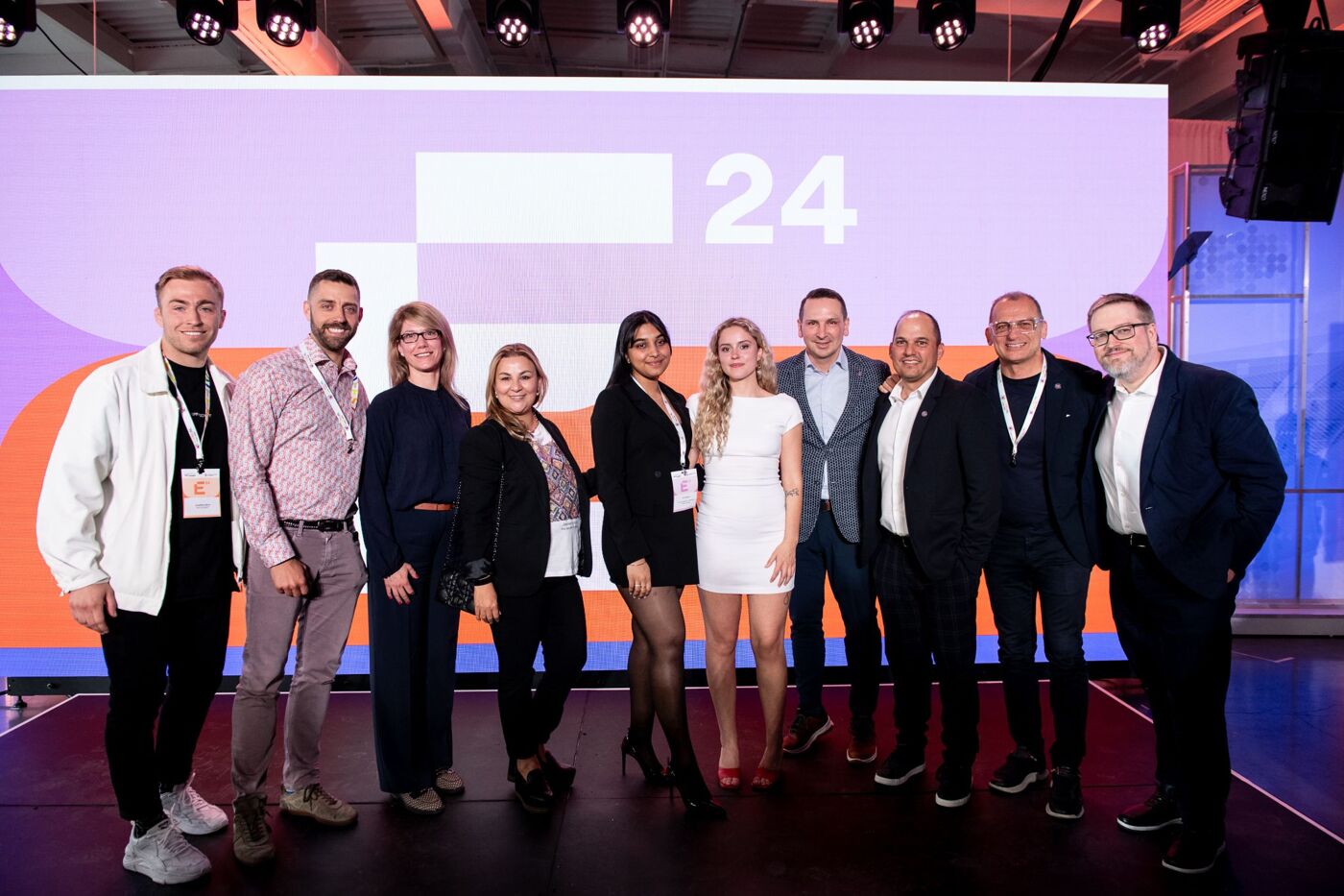 A group photo of twenty-four individuals, comprised of men and women in business casual attire, stands in front of a large screen displaying the number "24".