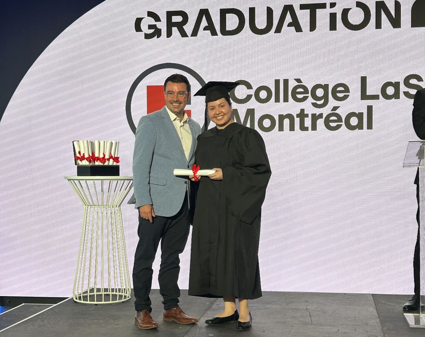 A graduating student in a black gown receives a diploma from a man in a suit, with the Collège LaSalle Montréal graduation backdrop visible.