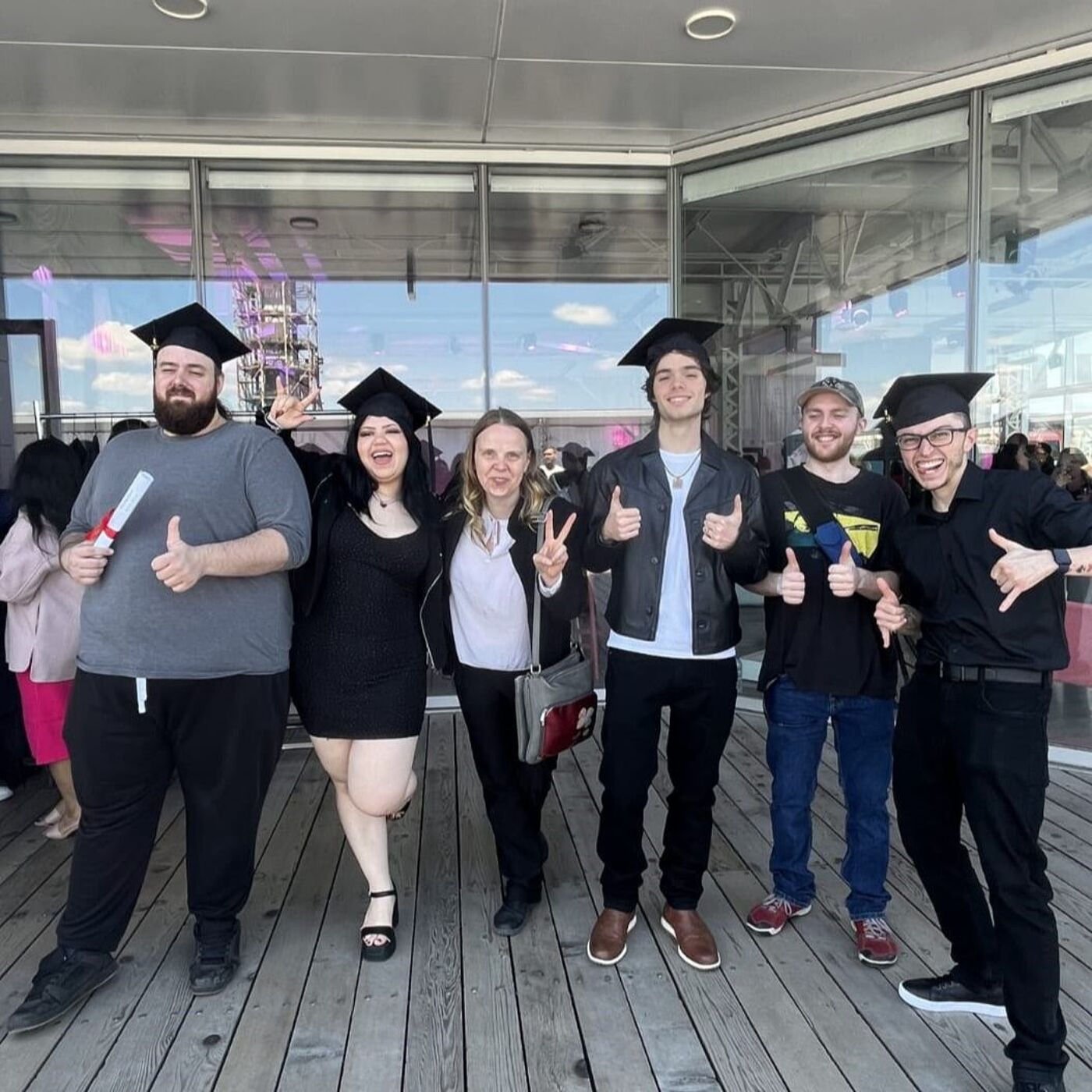 Seven graduates in caps and gowns pose for a photo on a boardwalk.