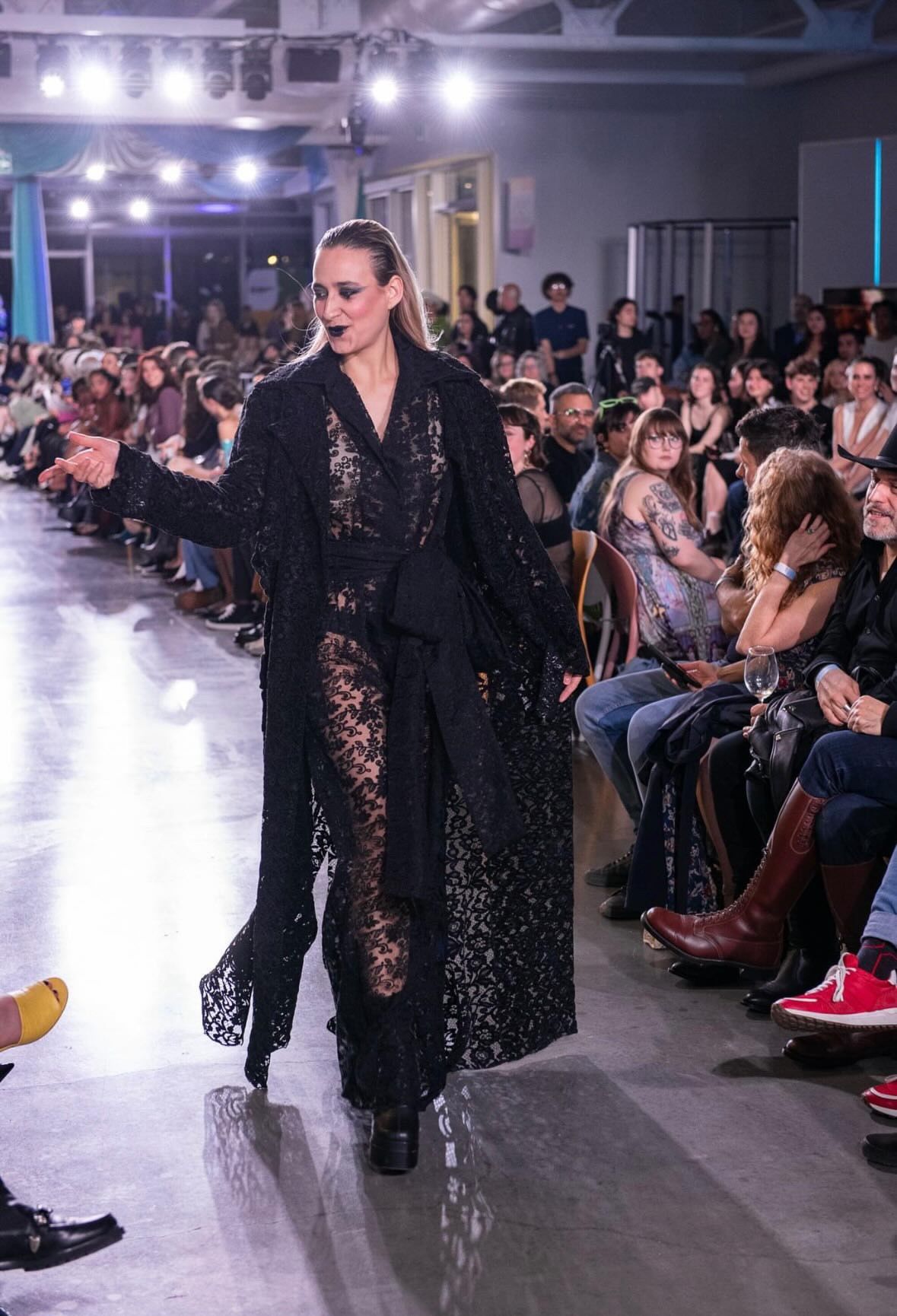 A model in a black lace dress walks down a runway during a fashion show, surrounded by a seated audience.