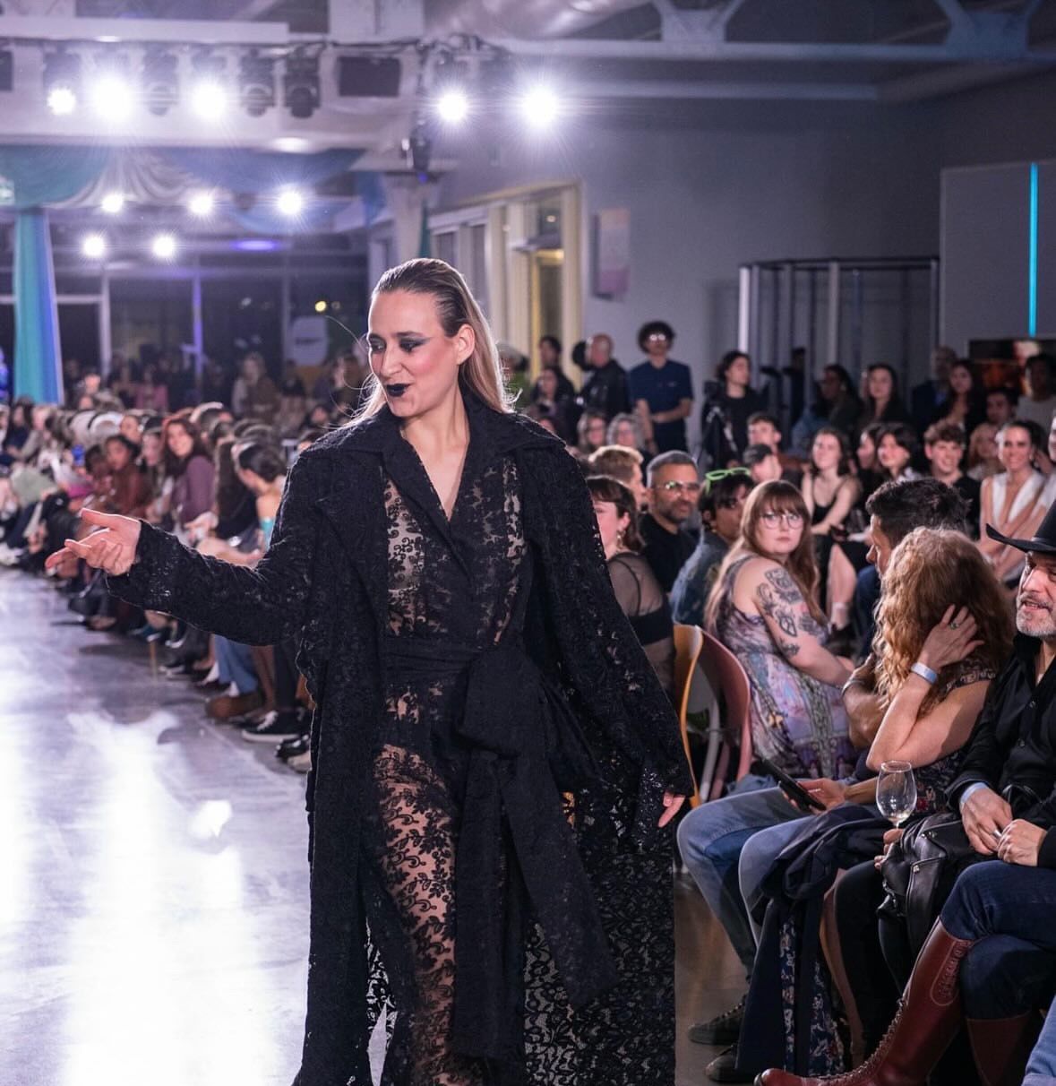 A model in a black lace dress walks down a runway during a fashion show, surrounded by a seated audience.