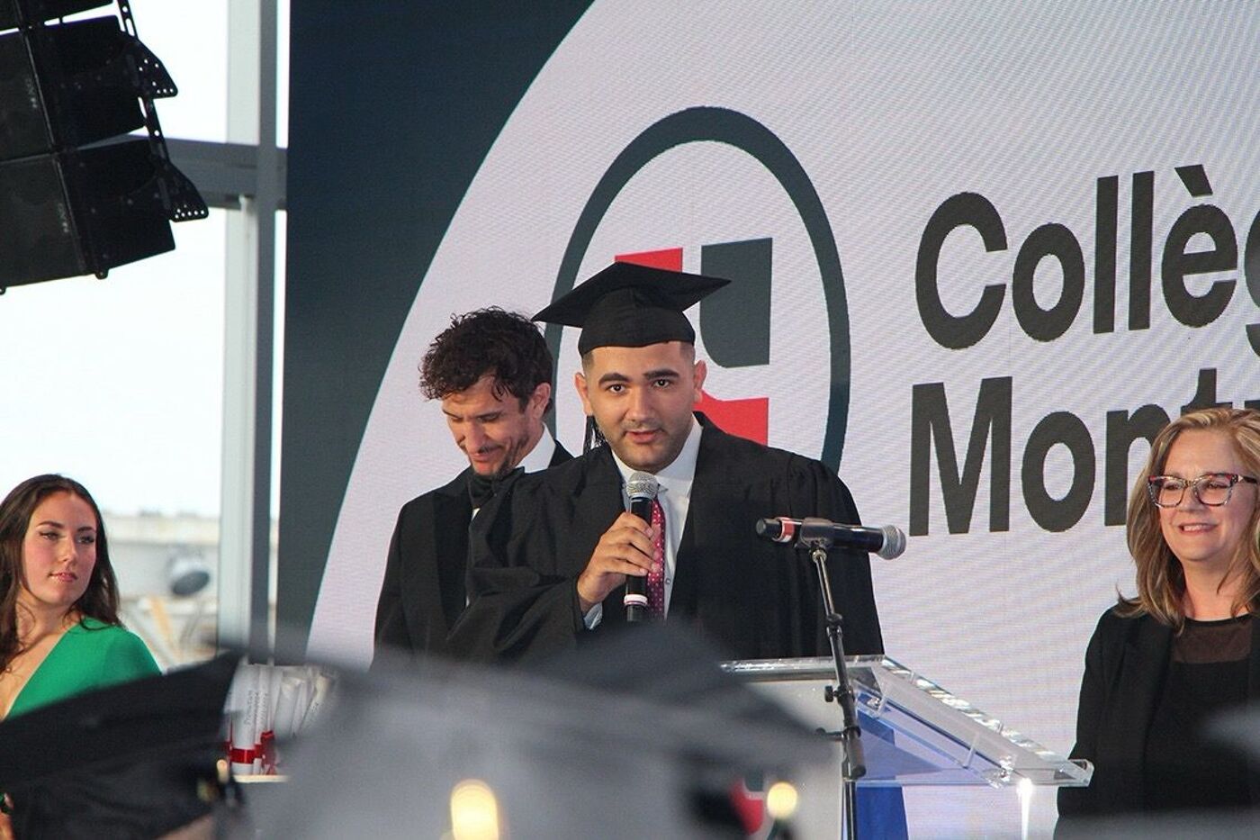 A graduate in a cap and gown stands at a podium giving a speech, flanked by another person in a suit and a woman in a dark suit, with a large college logo in the background.