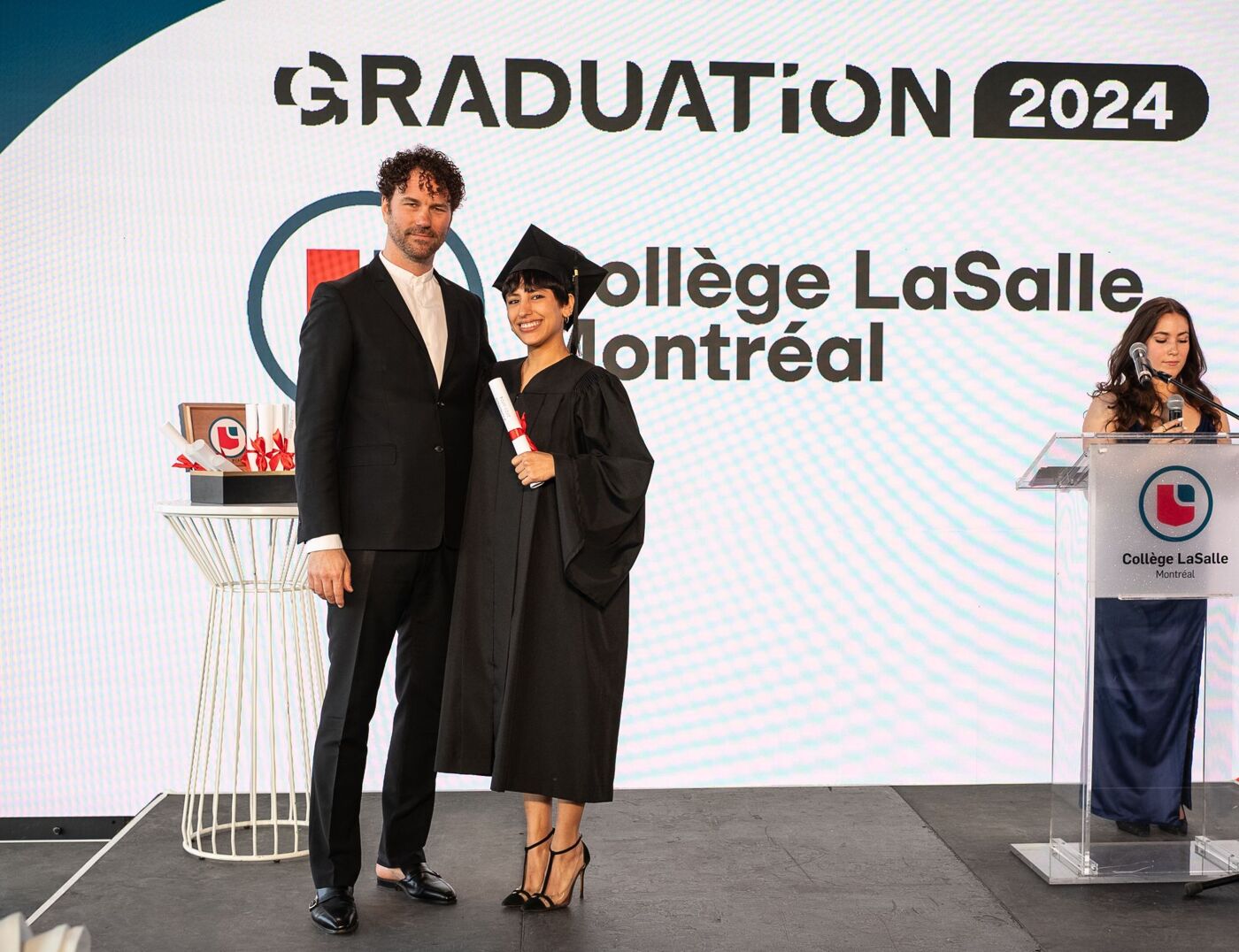 Two people in graduation attire stand next to a man in a suit, with a woman in a navy dress standing at a podium in the background, all in front of a banner that reads "GRADUATION 2024 Collège LaSalle Montréal".