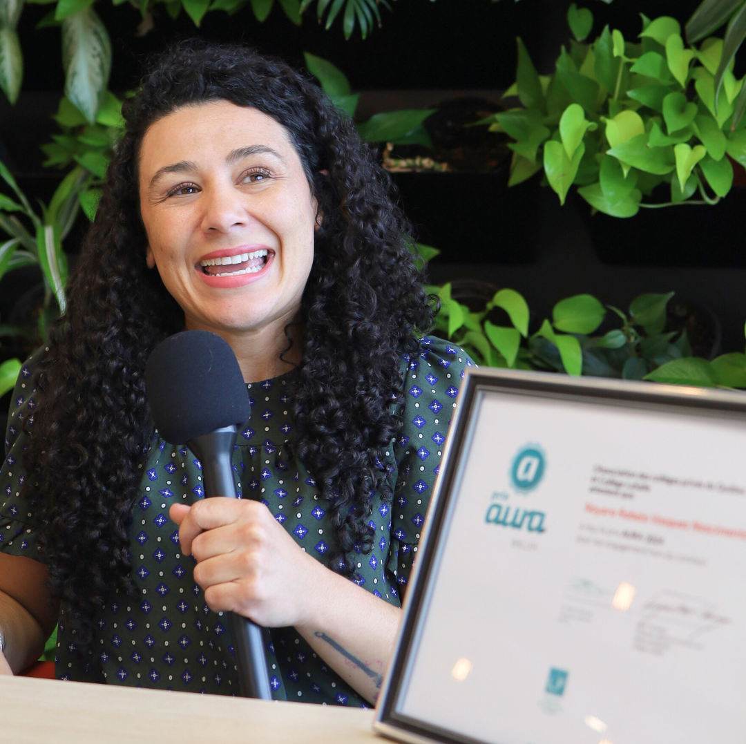 A woman with curly hair smiles while holding a microphone in front of a wall of plants, with a framed award visible on the table in front of her.