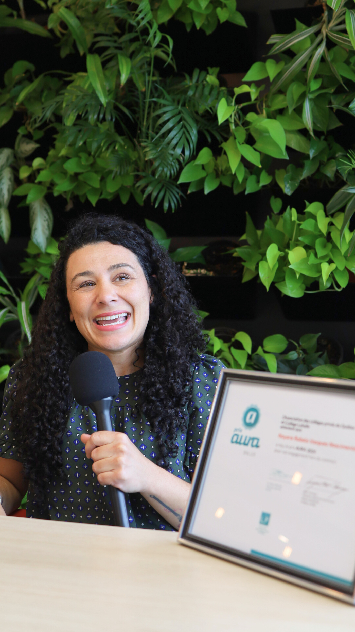 A woman with curly hair smiles while holding a microphone in front of a wall of plants and a framed certificate.