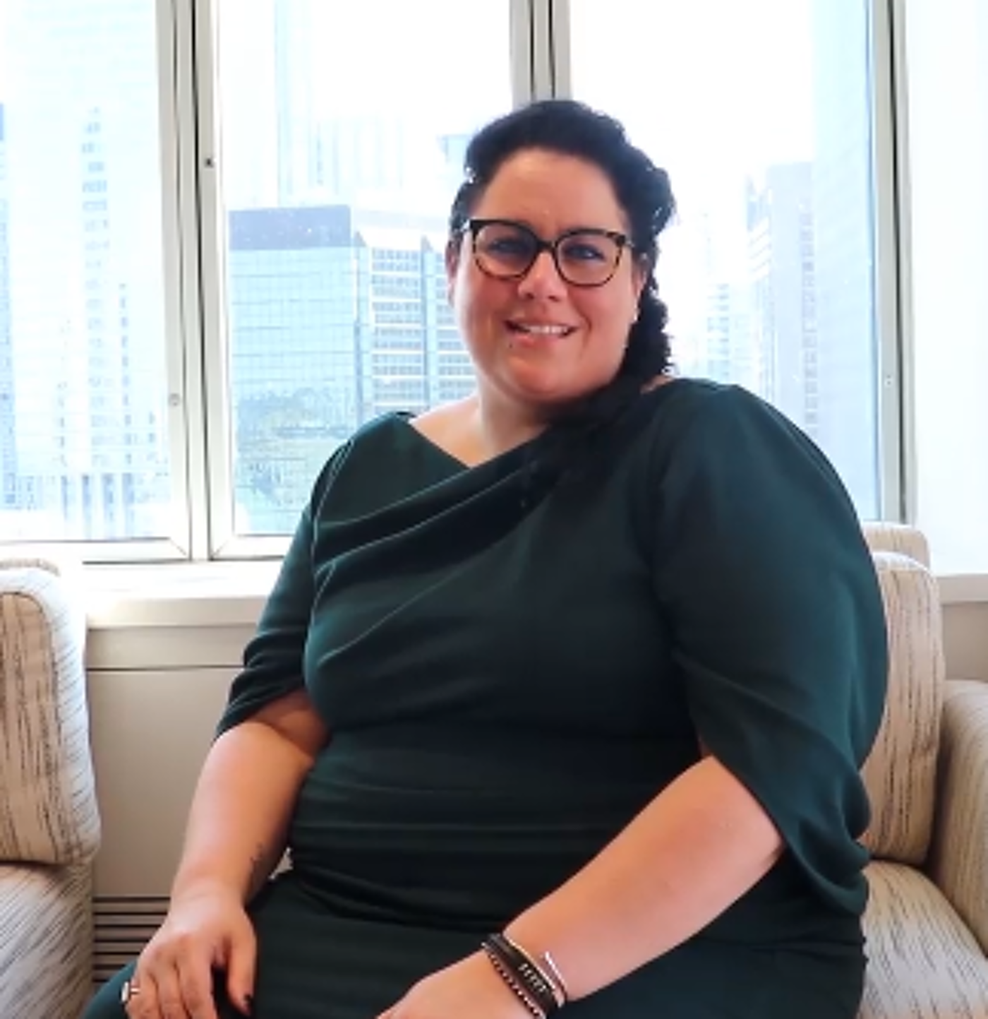 A plus-size woman with dark hair and glasses smiles while seated in a chair in front of a window overlooking a cityscape, wearing a dark green dress.