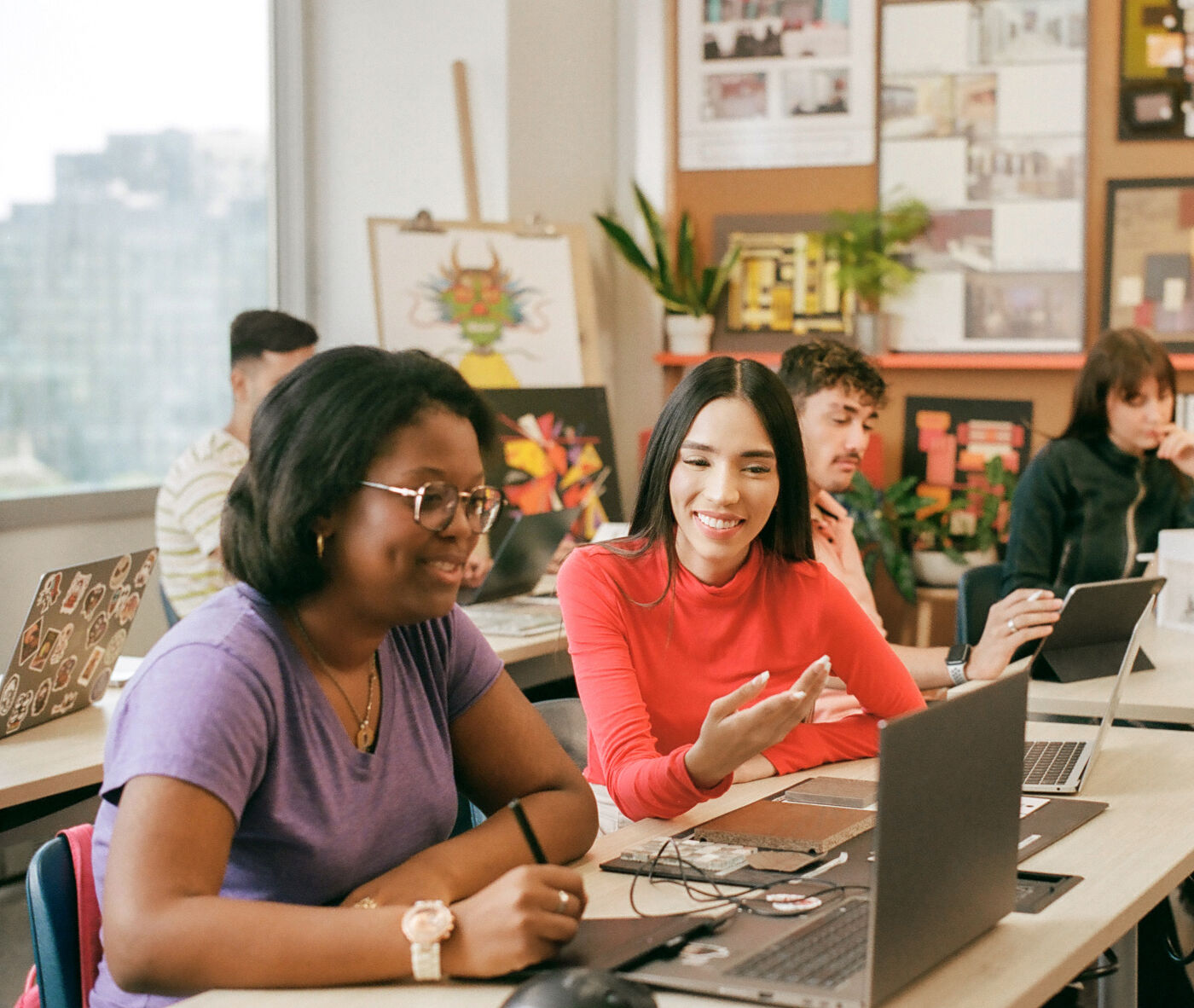 Adult students of various backgrounds engage with laptops and tablets in a bright classroom.