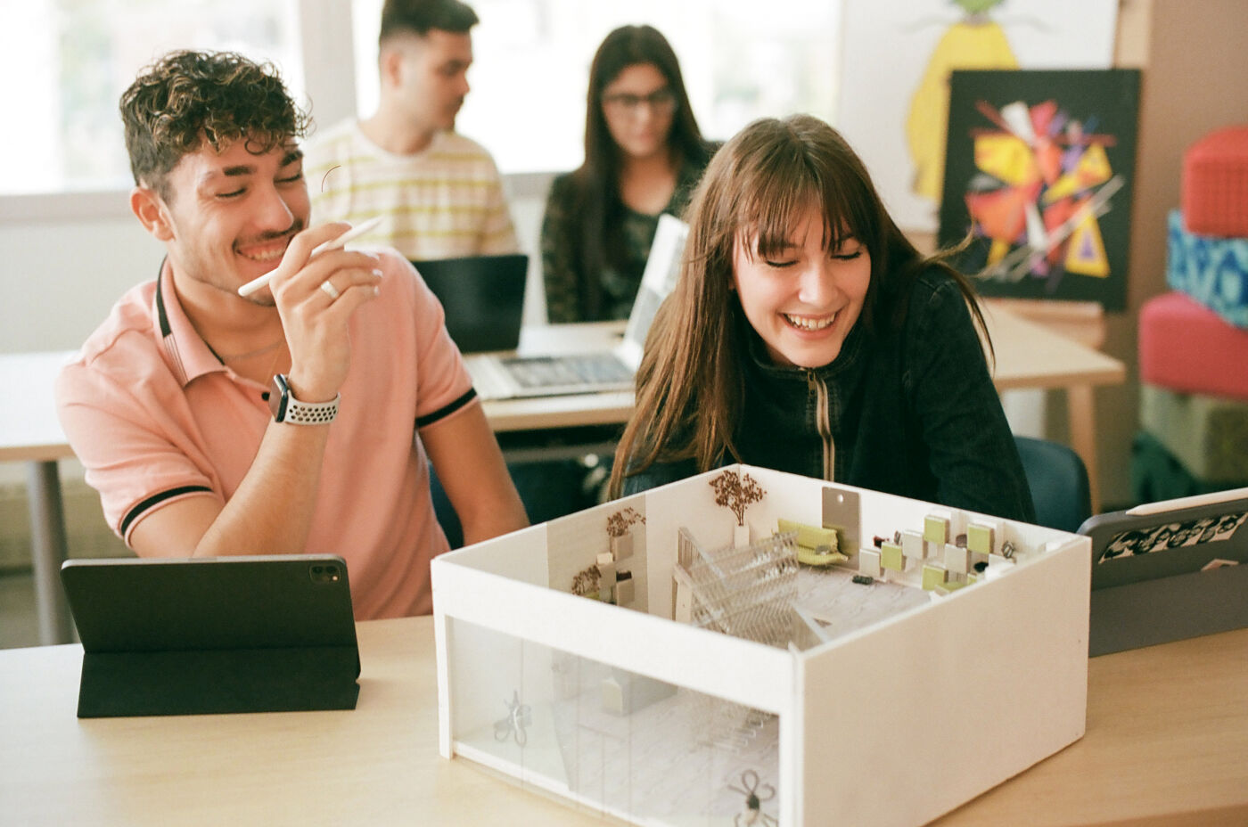 Two students laugh while examining a detailed architectural model in a classroom setting.