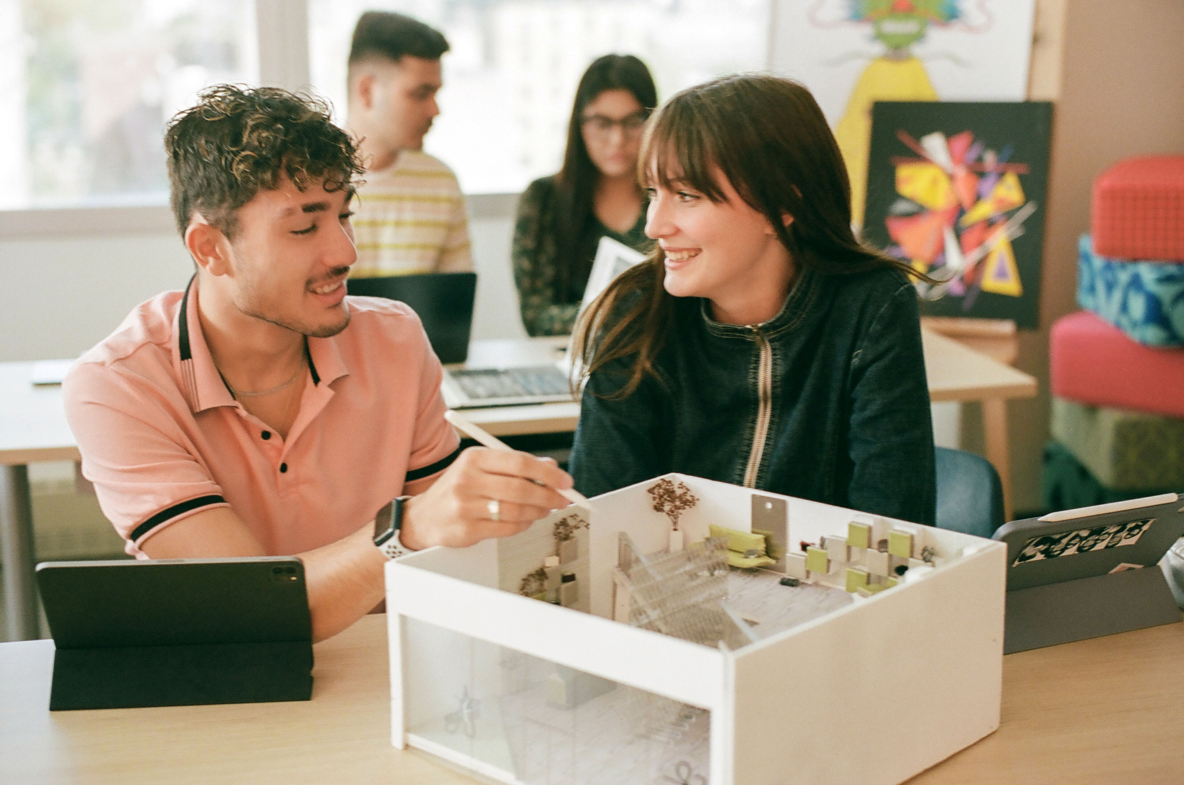 Two students laugh while examining a detailed architectural model in a classroom setting.