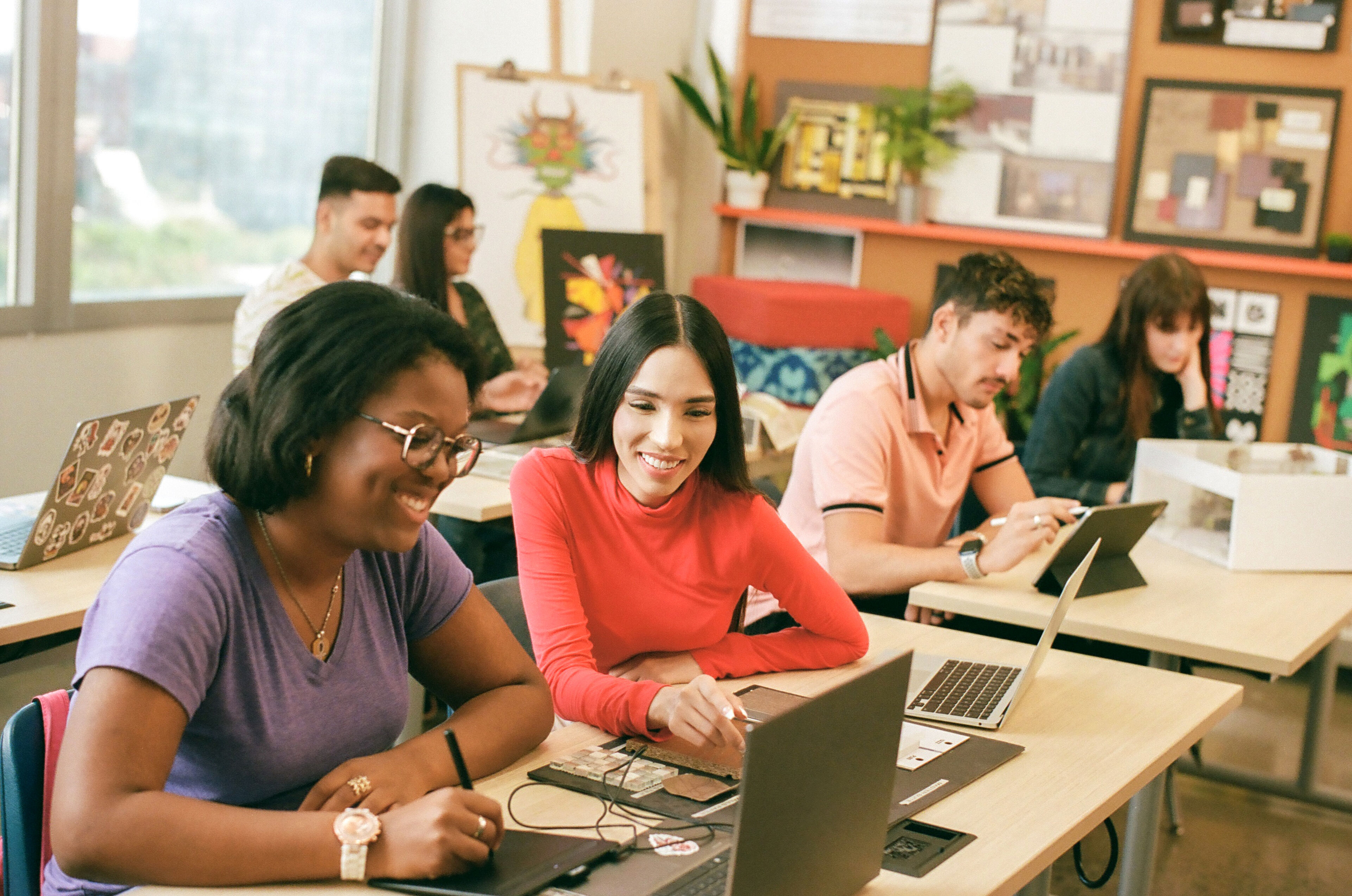 Adult students of various backgrounds engage with laptops and tablets in a bright classroom.