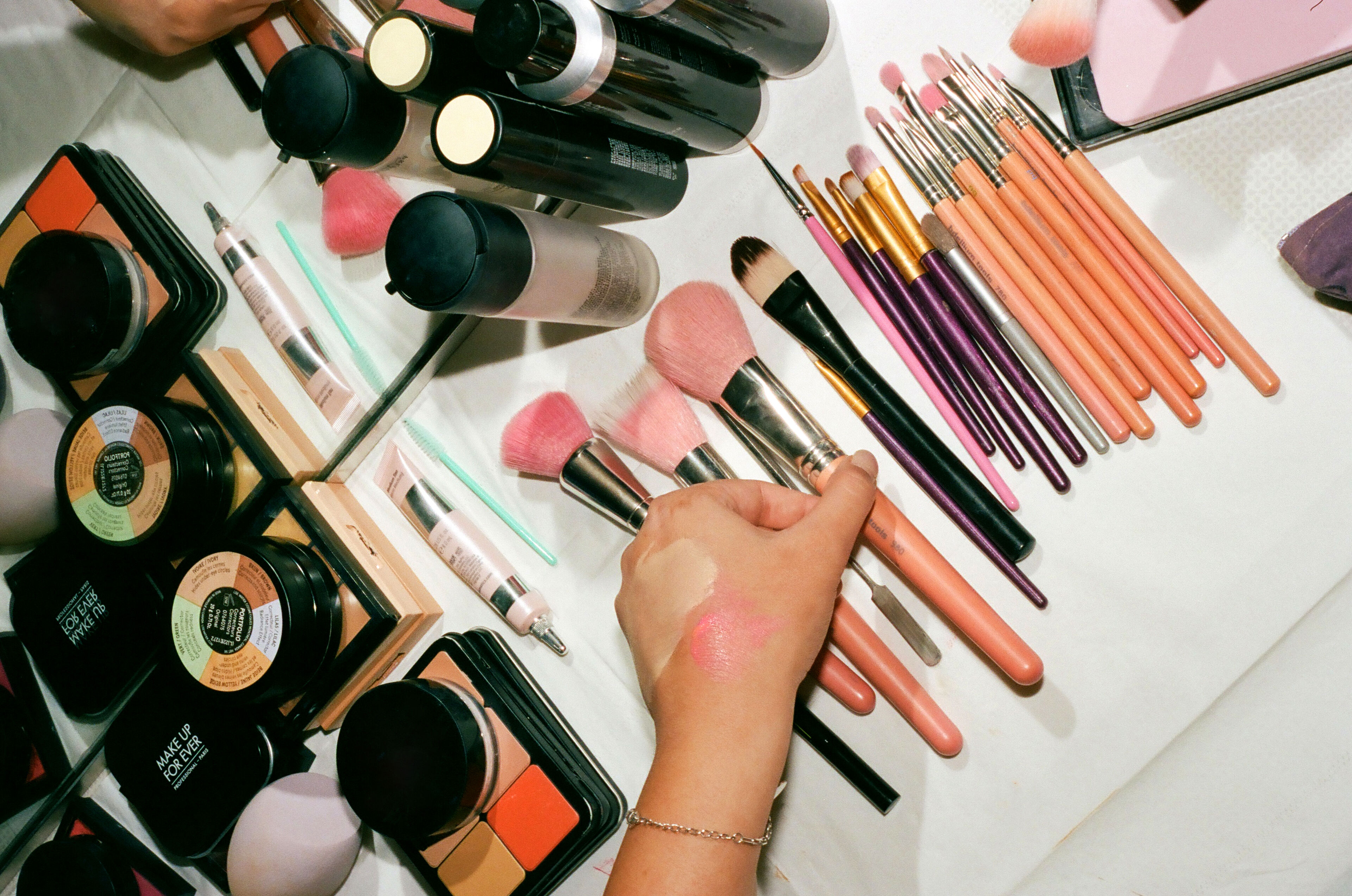 A person's hand with swatched makeup surrounded by various cosmetics and applicator brushes on a white surface.