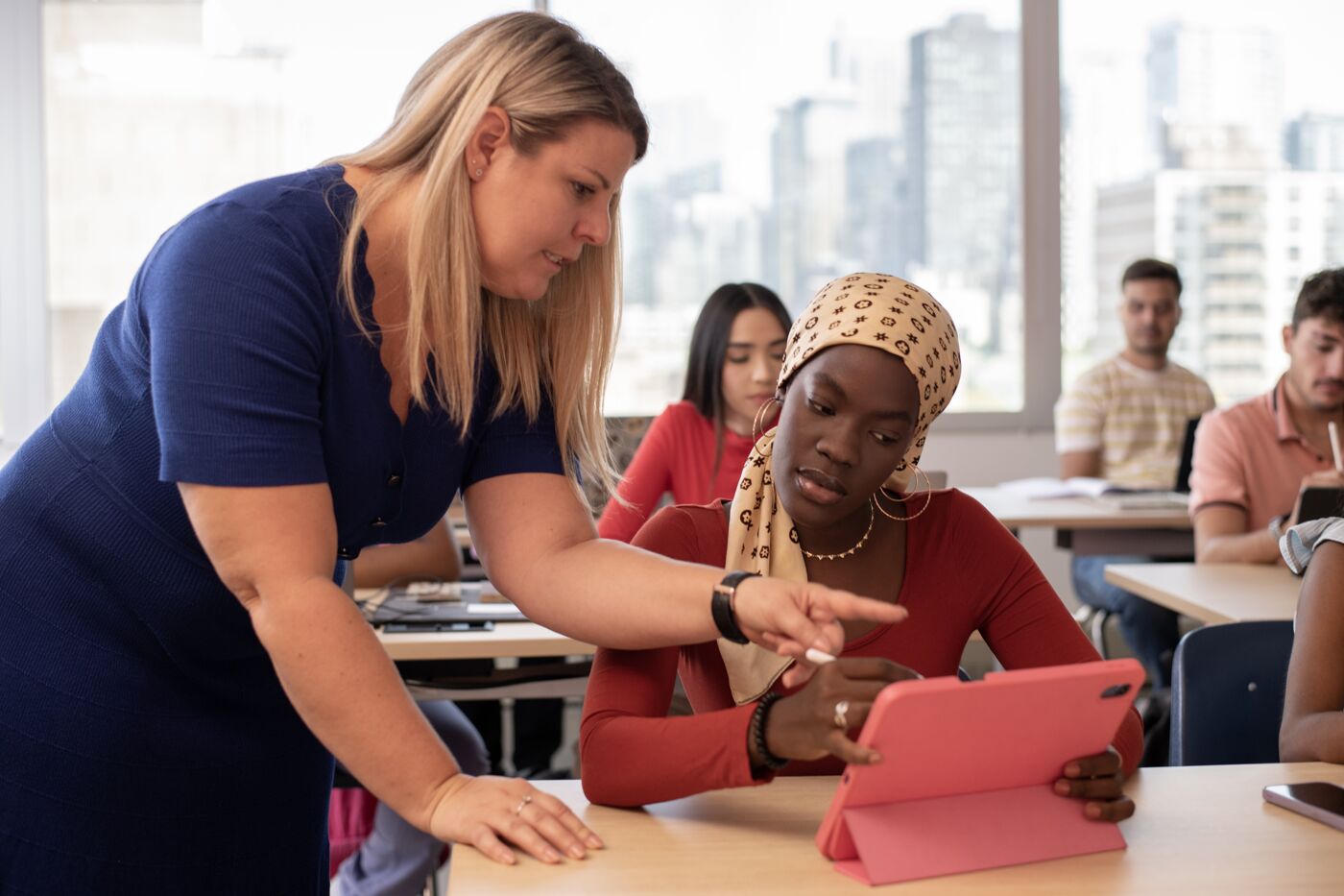 A teacher assists a student with a tablet in a classroom setting, with other students focused on their work in the background.