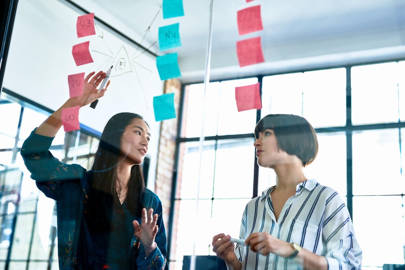 Two women collaborating in an office setting, using a glass board with sticky notes for a brainstorming session.