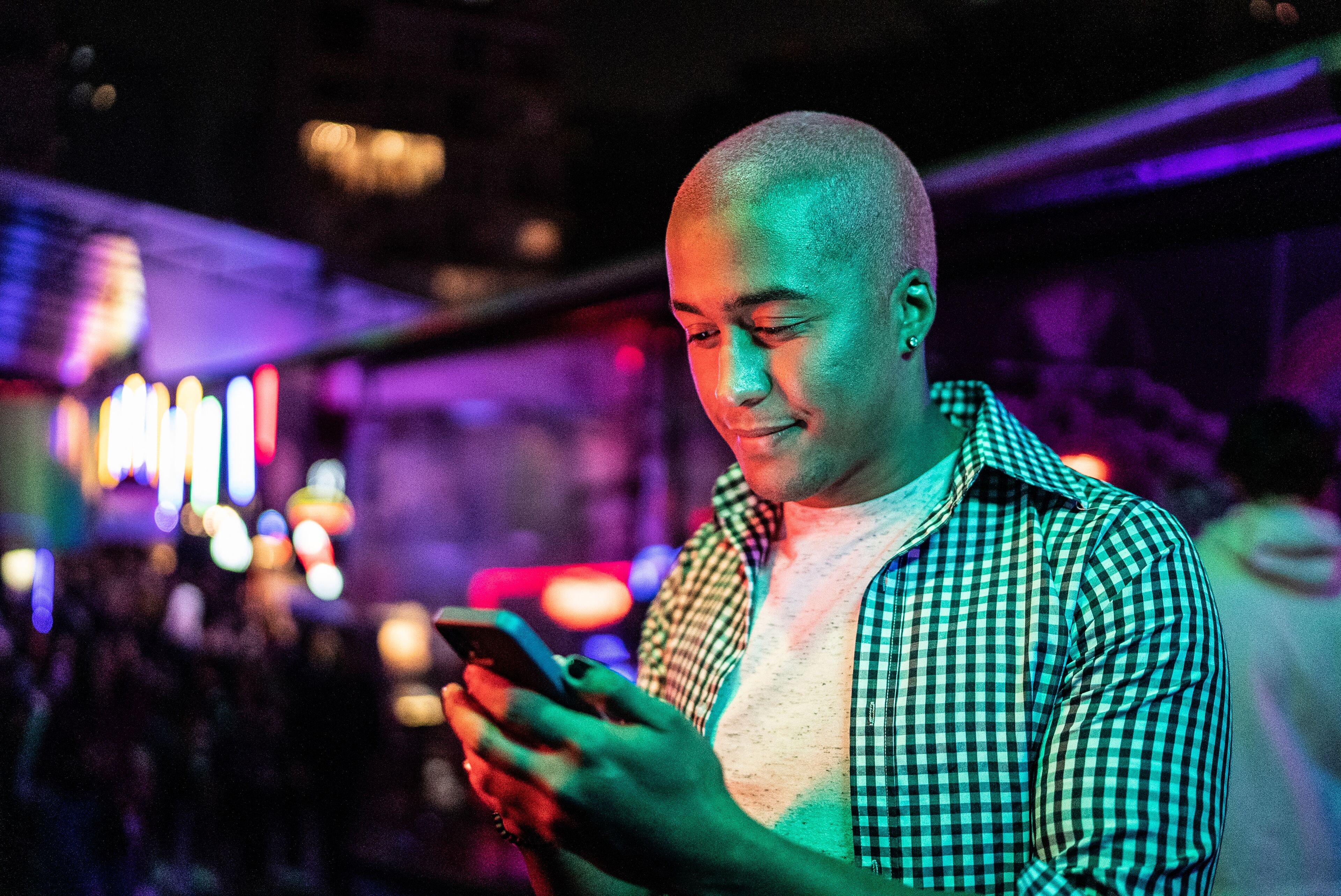 A bald man in a checkered shirt smiling at his phone amidst colorful neon lights.