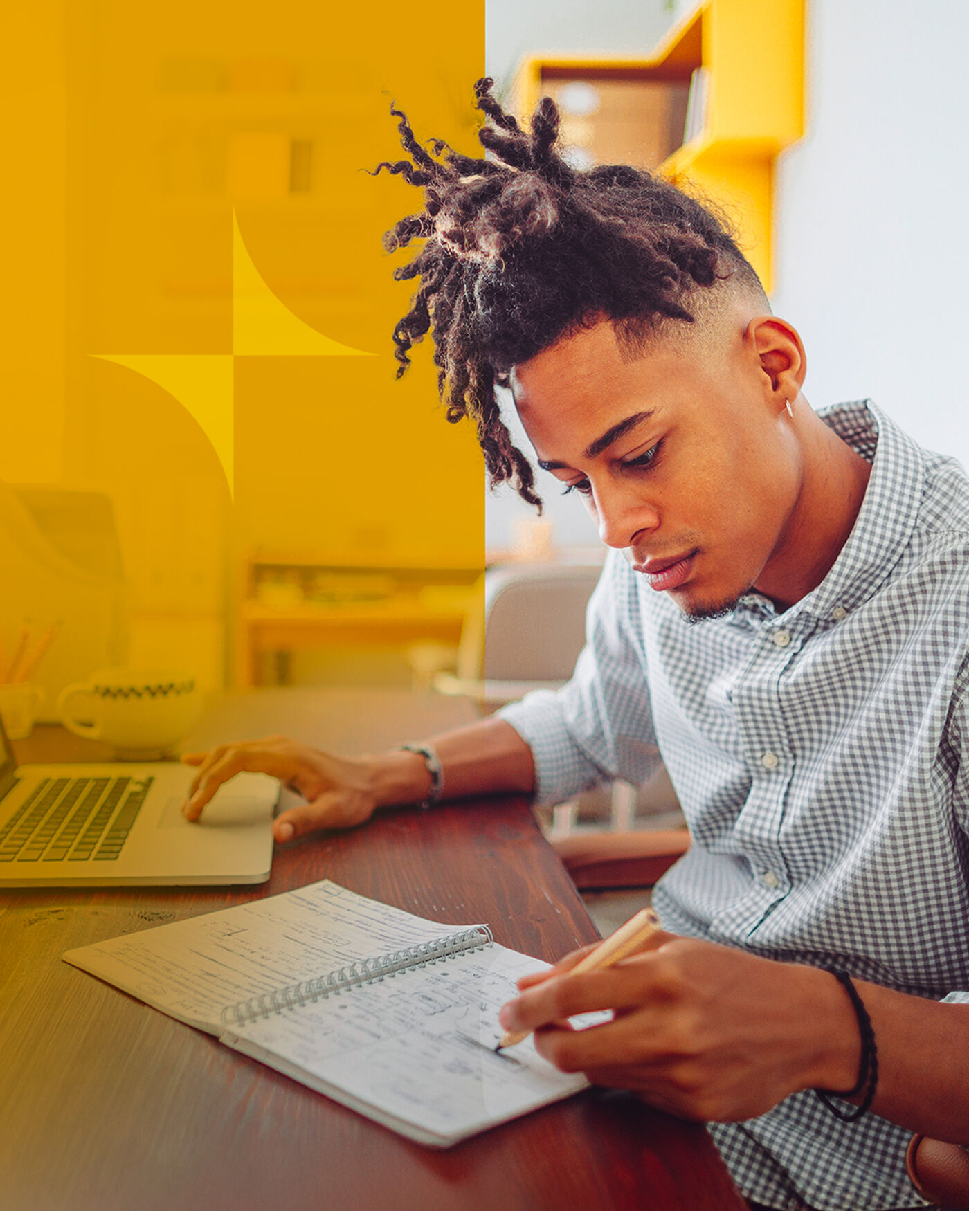A young man with dreadlocks is sitting at a desk, focused on his work. He is using a laptop with one hand and writing in a notebook with the other, suggesting he is studying or working on a project.