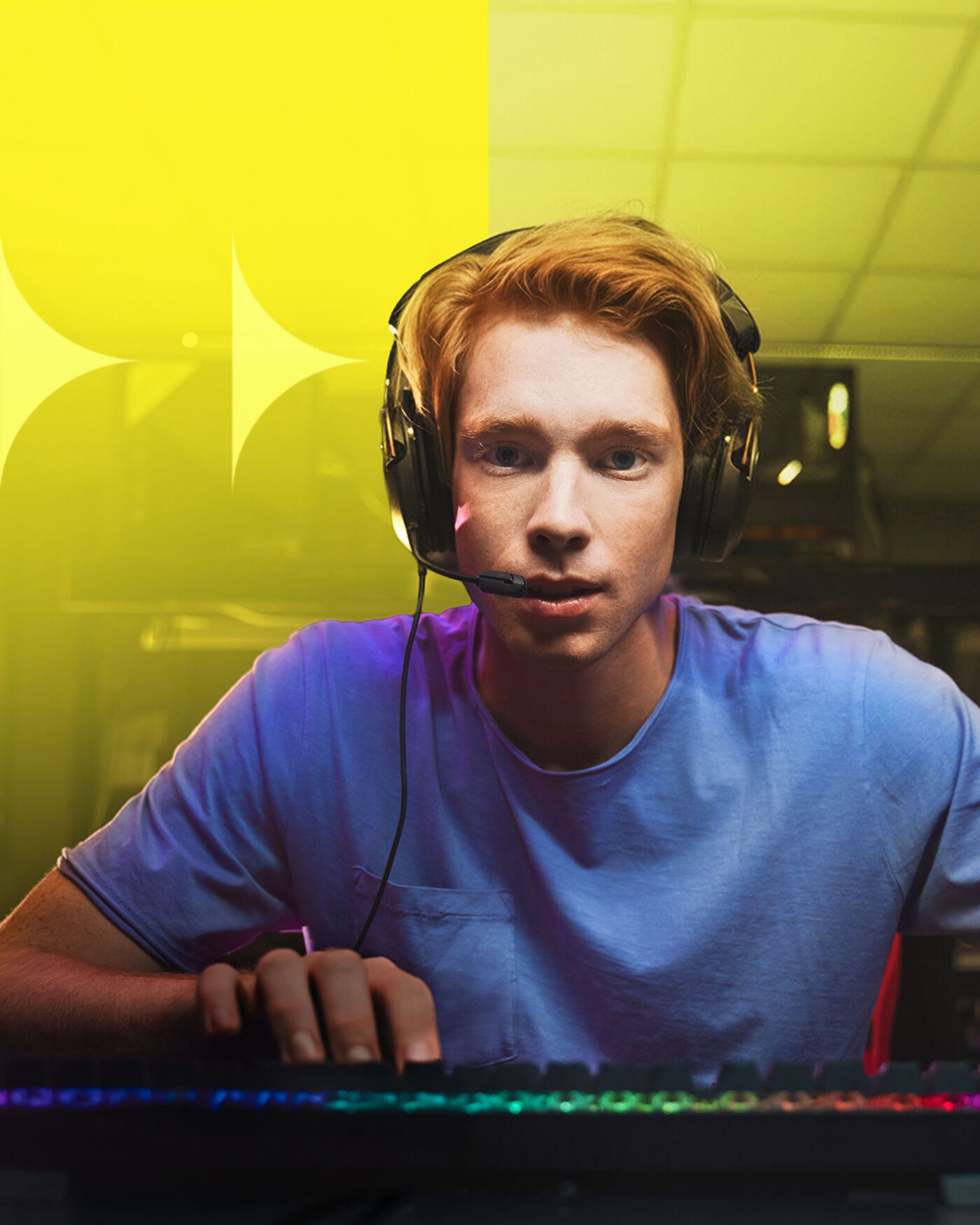 A young man with red hair is wearing a headset and a blue t-shirt while playing a video game. The keyboard in front of him has rainbow-colored lights. The background is a mix of yellow and dark colors.