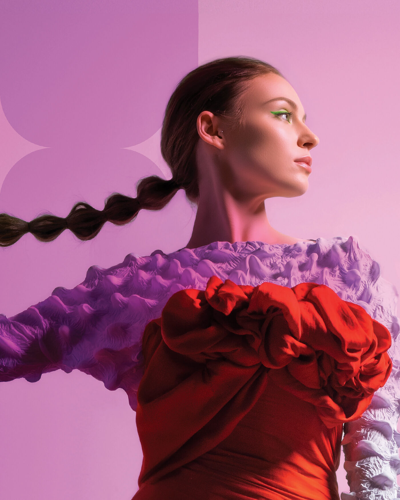 A woman with fair skin and brown hair styled in a thick braid poses in front of a pink background. She is wearing a textured lavender top with long sleeves and a large red fabric embellishment on the front.