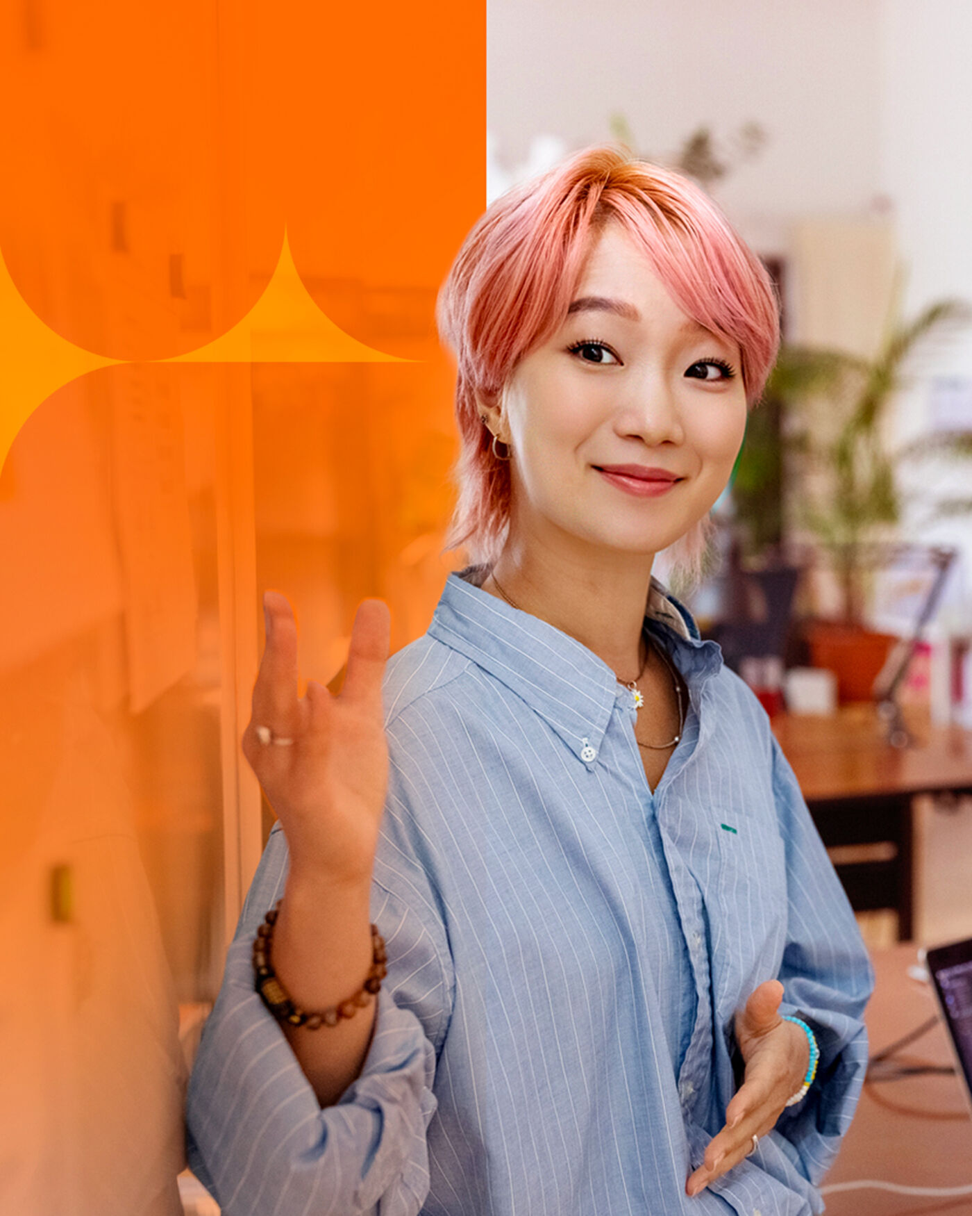 The image shows a smiling woman with pink hair, wearing a light blue striped shirt and bracelets. She is standing in an office setting with a desk and plants visible in the background.