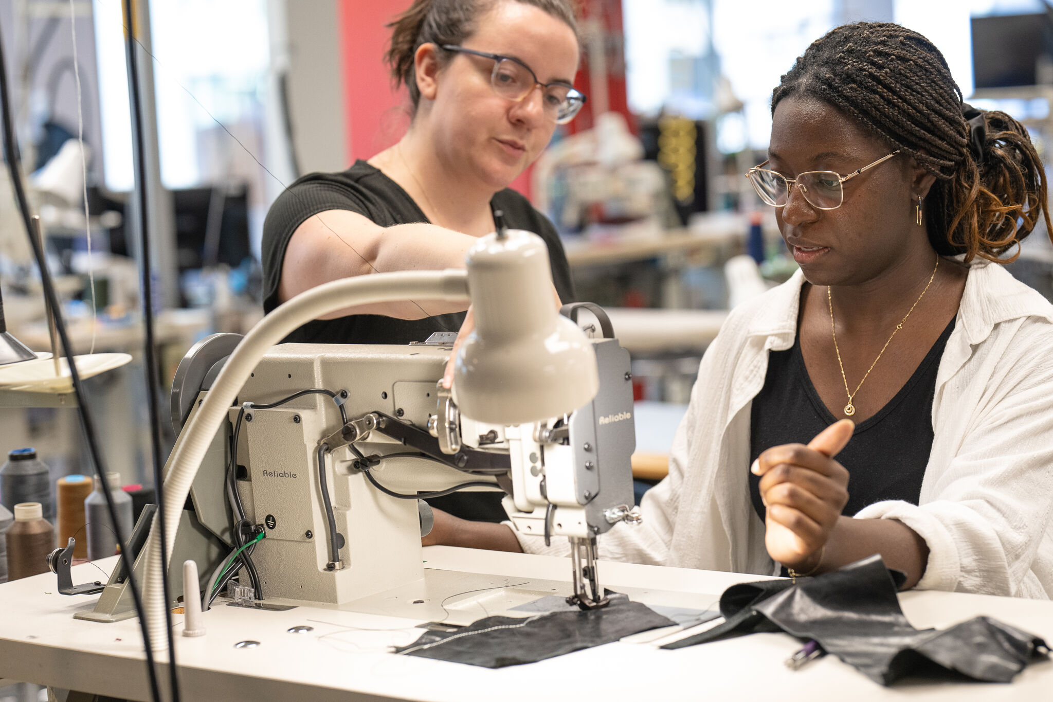 Two women focus intently on sewing fabric with an industrial sewing machine in a workshop setting.