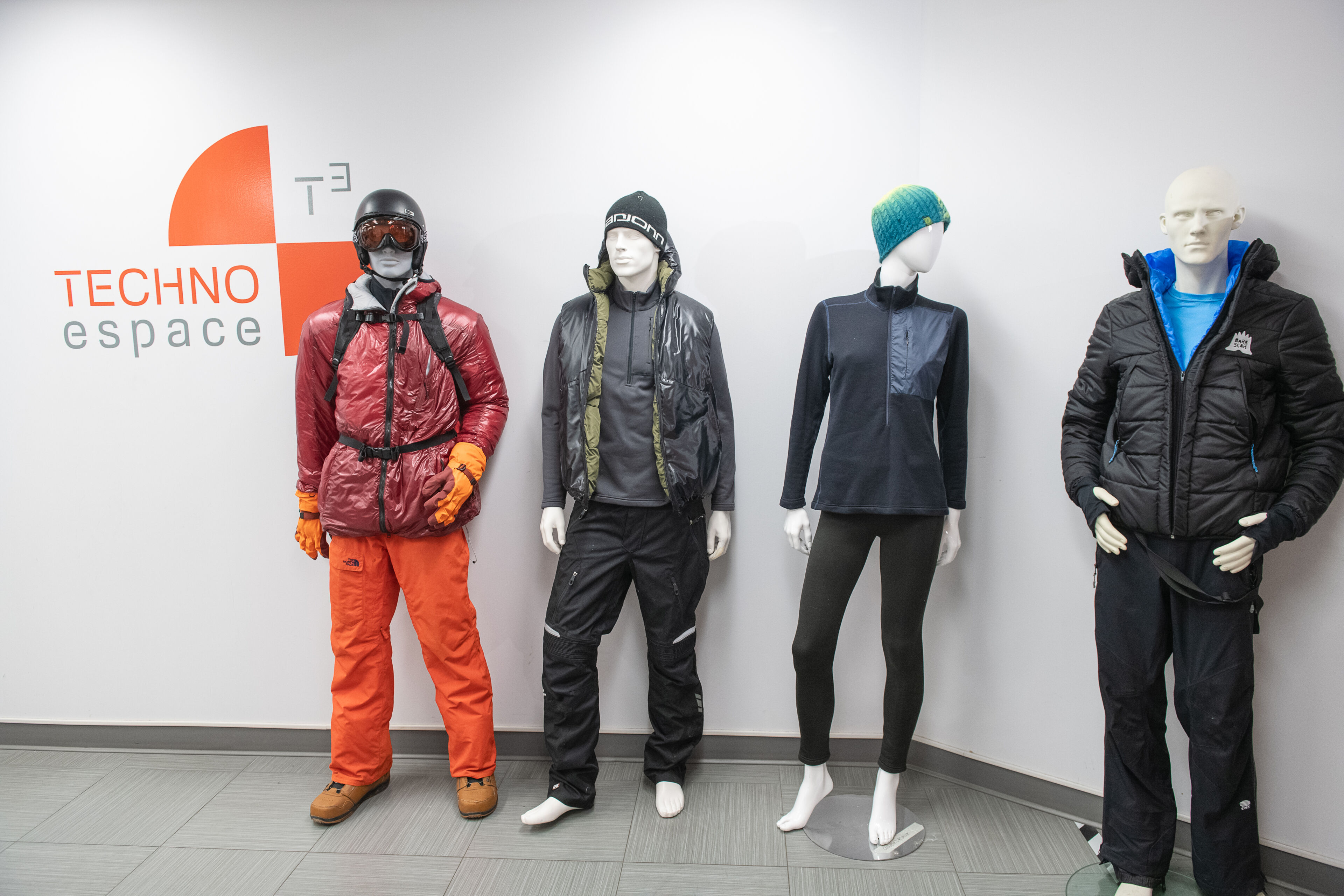 Four mannequins dressed in winter sportswear, including insulated jackets and pants, displayed beside a Techno Espace sign.